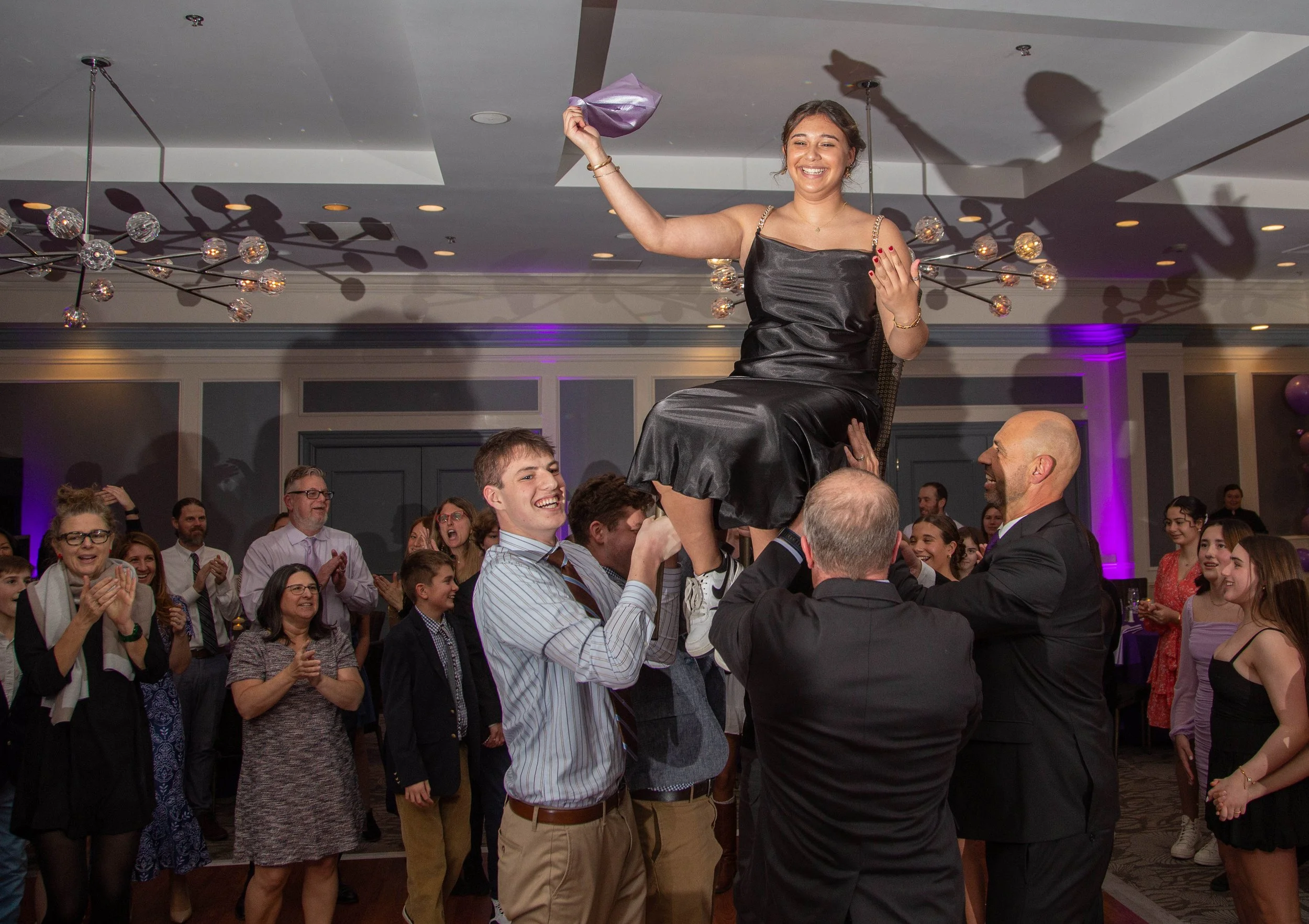 A woman in a black dress being lifted in a chair by several men during a celebration or wedding reception, with guests clapping and smiling in the background.