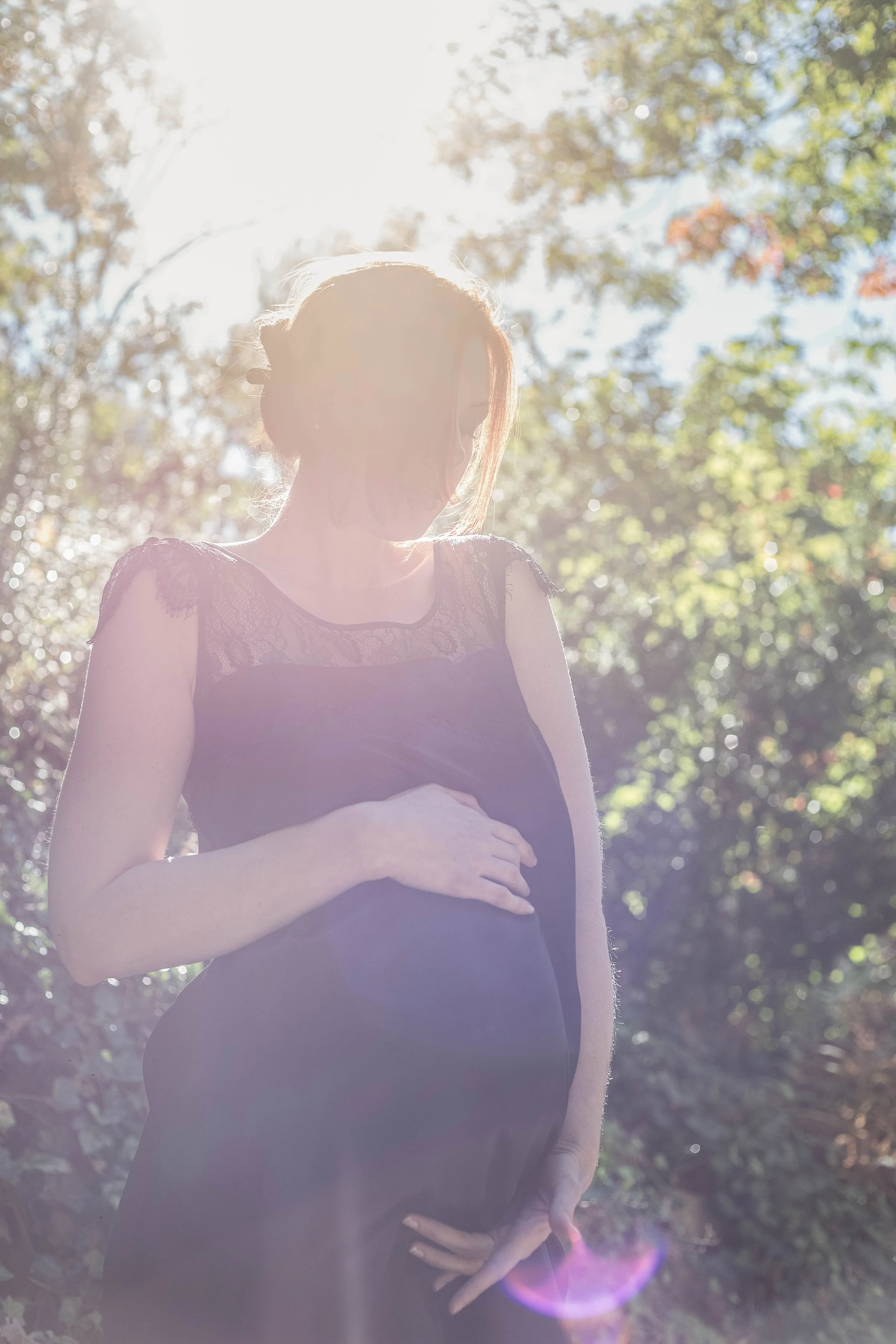 A pregnant woman in a black dress standing outdoors with sunlight behind her, holding her belly.