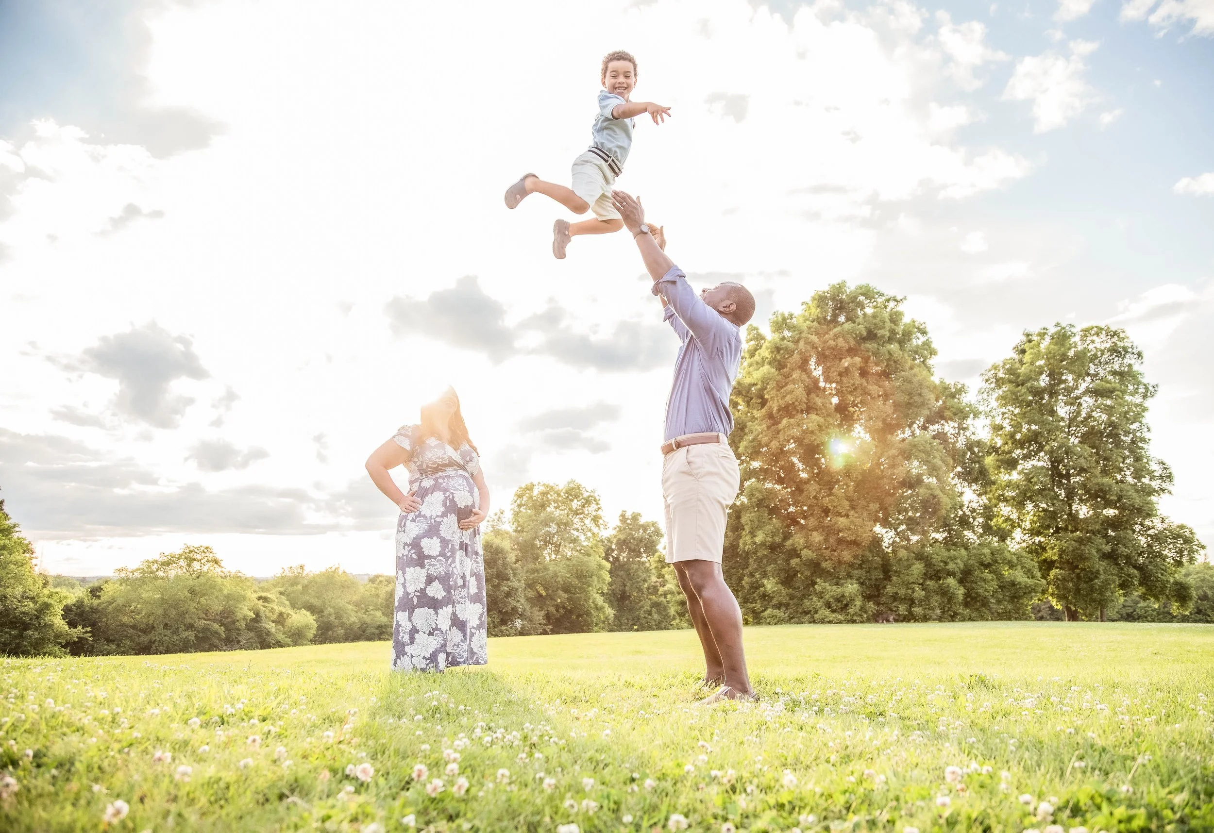 A man lifting a young boy into the air on a grassy field, with a woman standing nearby, all outdoors on a sunny day with clouds and trees in the background.