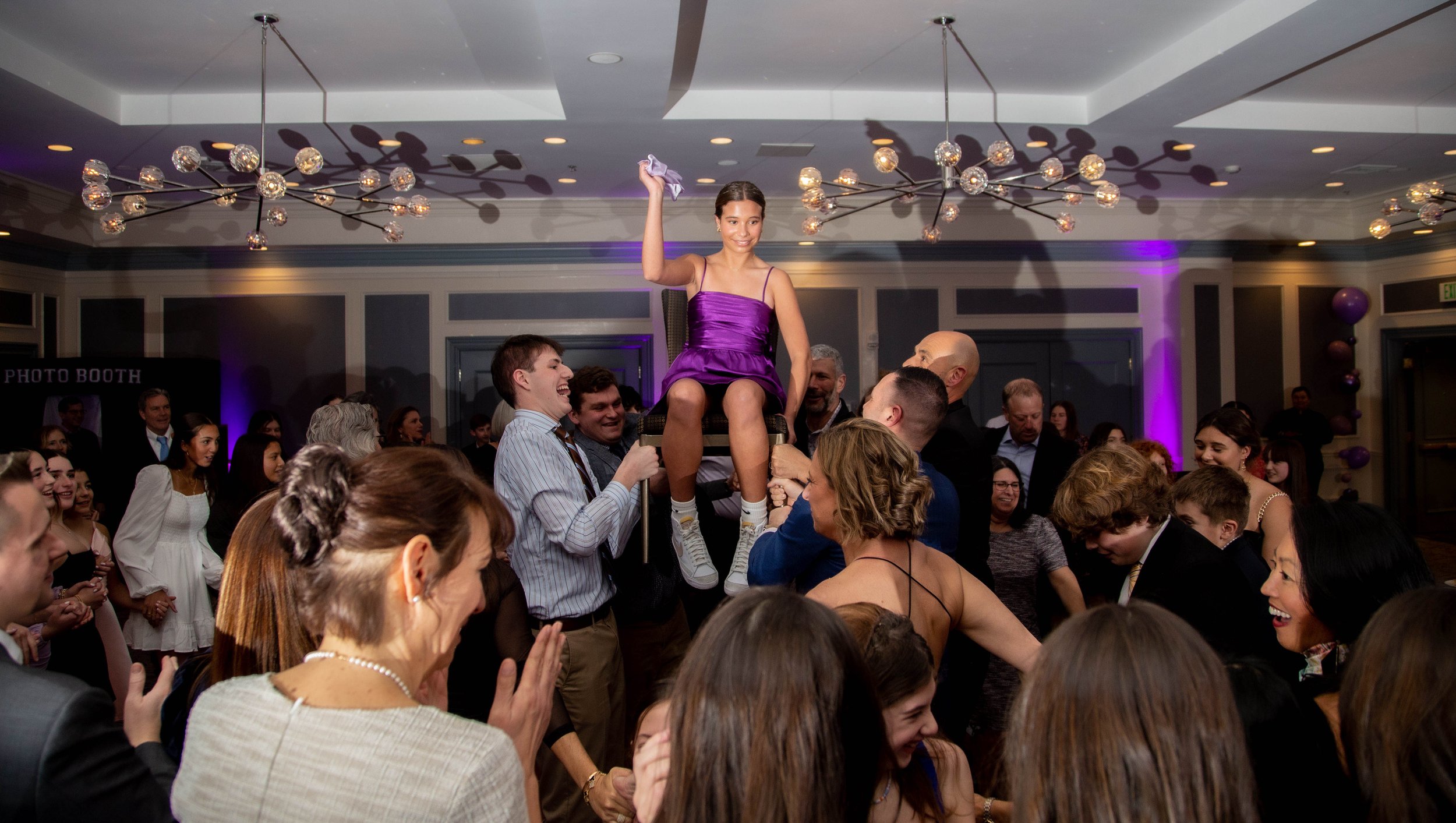 A group of people celebrating at a party, with a young woman in a purple dress sitting on a chair being lifted by others, smiling and holding a purple cloth in her raised hand.
