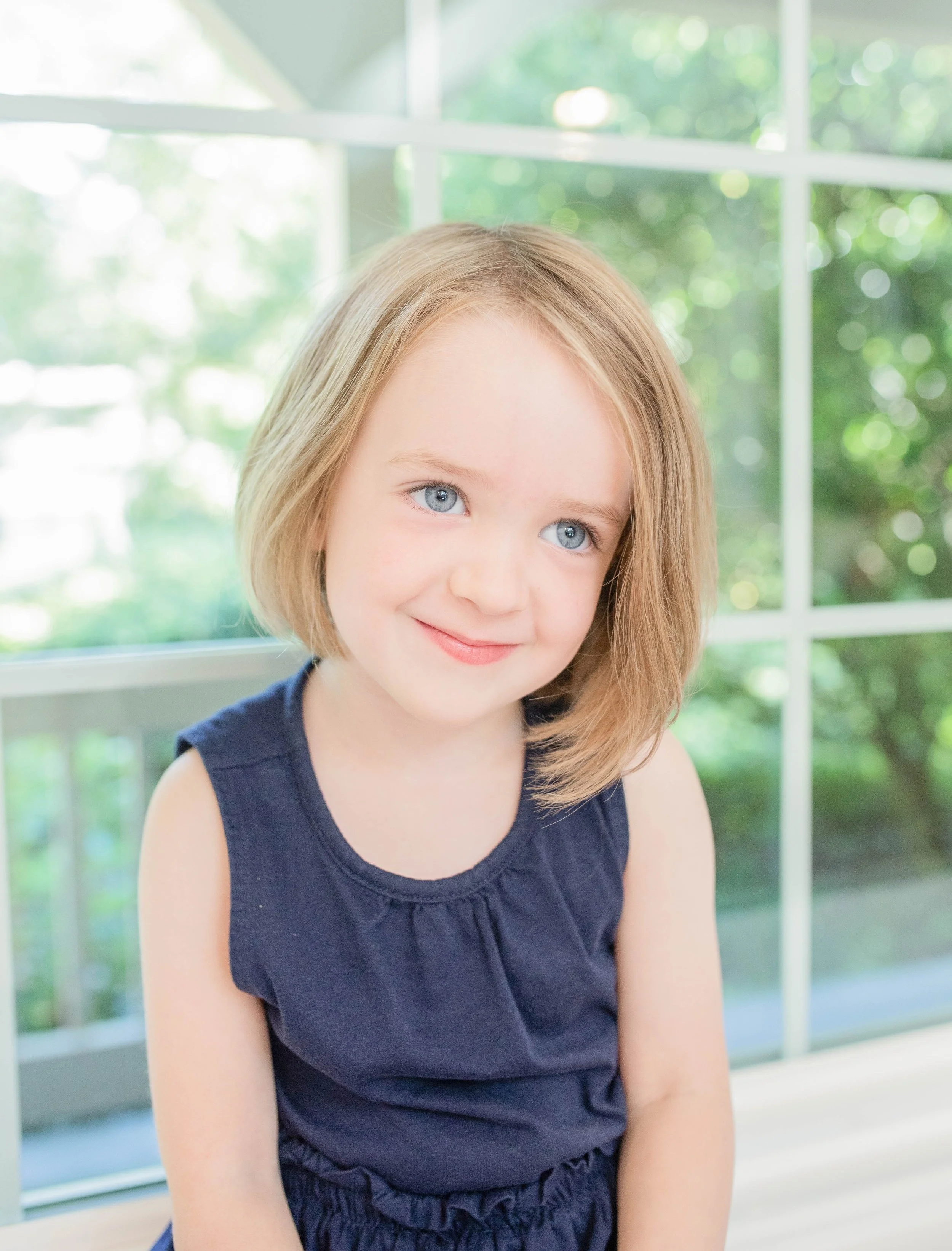 A young girl with blonde hair and blue eyes wearing a navy blue dress sitting in front of a window with greenery outside.