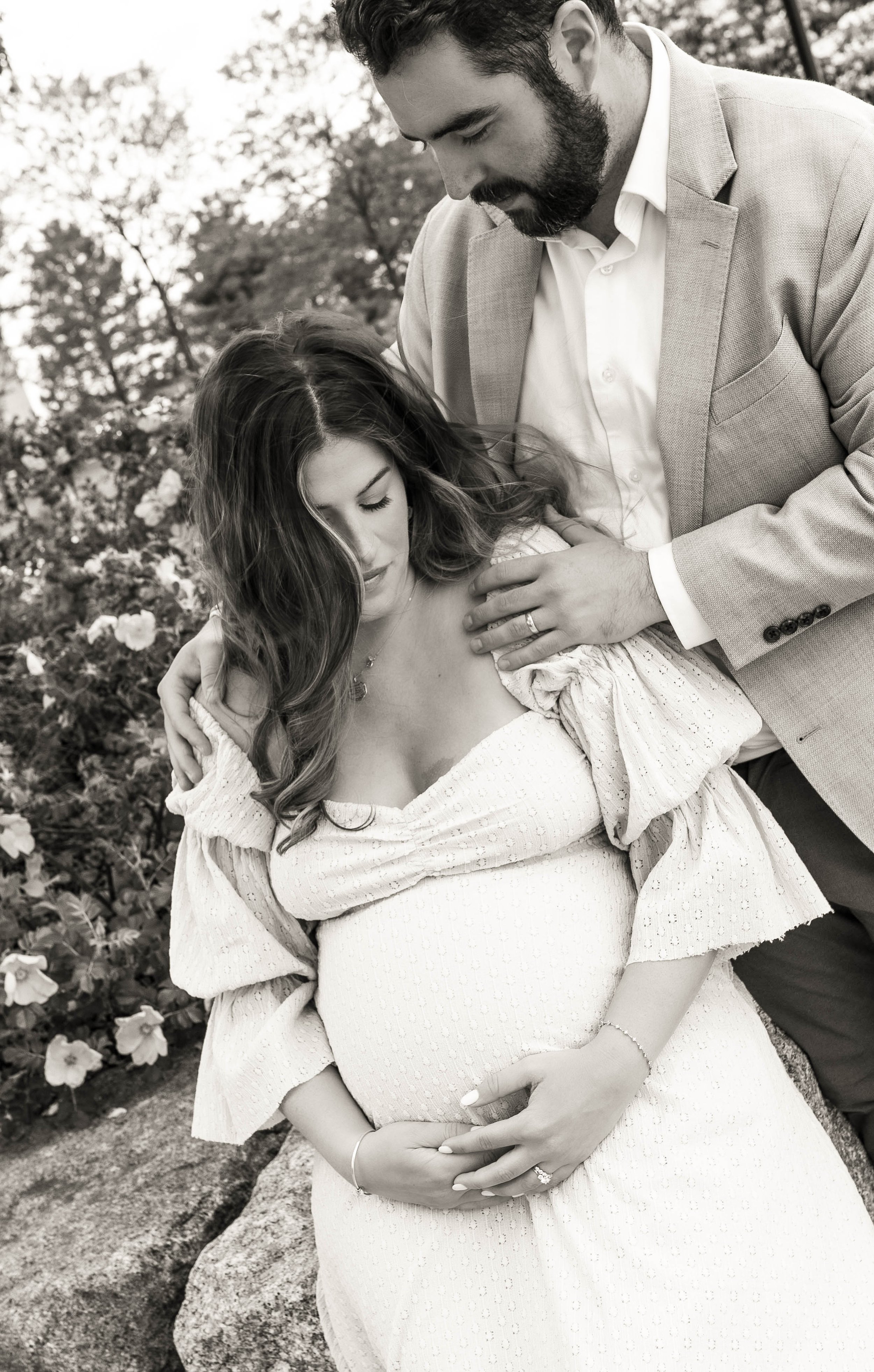 A black-and-white photo of a pregnant woman with long wavy hair in a white dress, holding her belly, and a man in a light suit with a beard, gently touching her shoulder in an outdoor setting with flowers and trees.