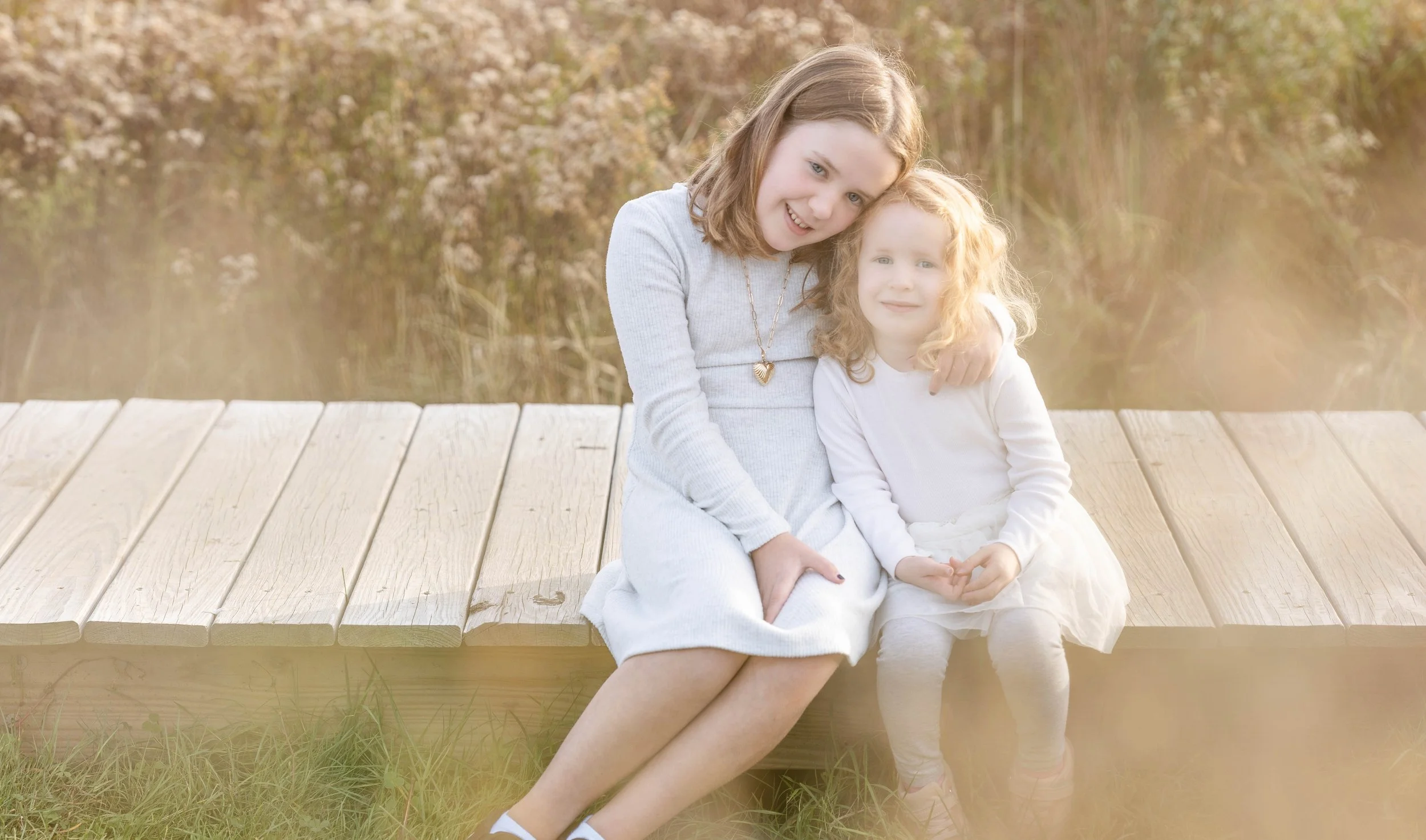 Two young girls sitting on a wooden dock outdoors, with the older girl hugging the younger girl, surrounded by blurred natural scenery and warm lighting.