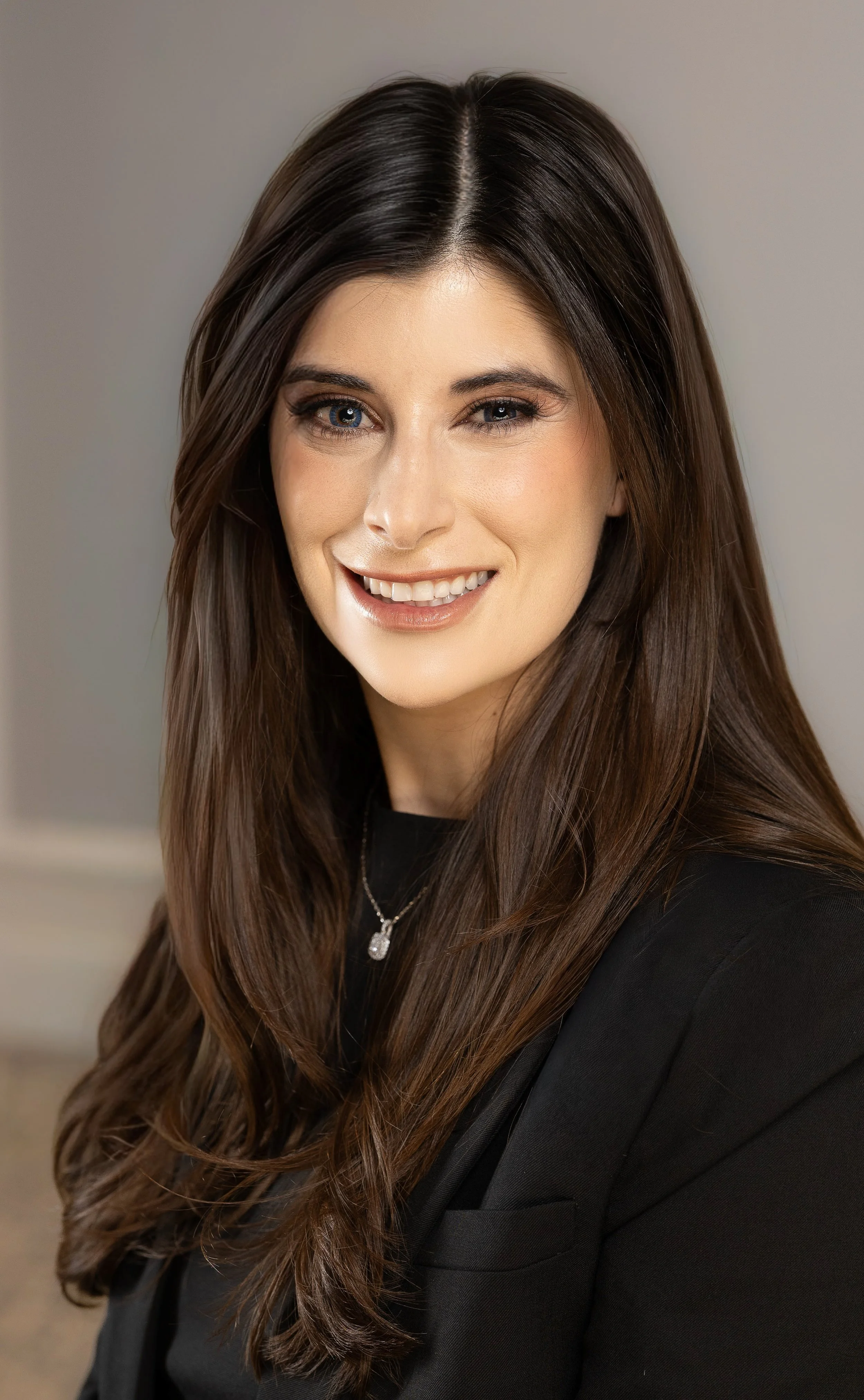 A young woman with long brown hair, gray eyes, and a smile, wearing a black blazer and a silver necklace, against a neutral background.