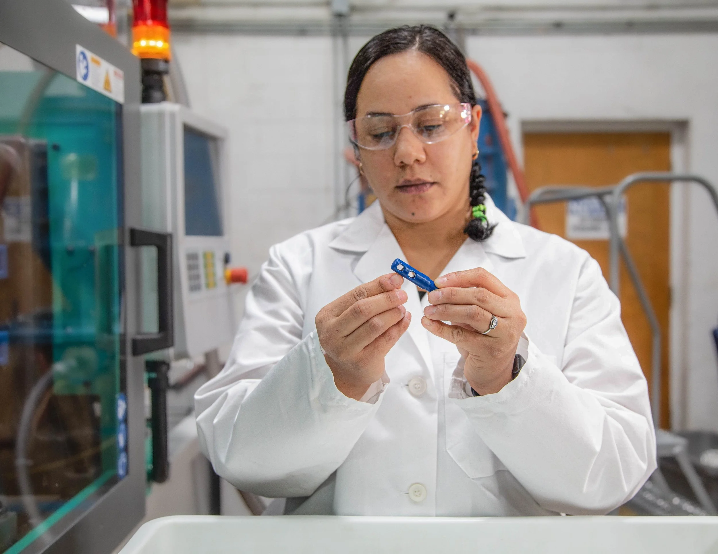 A woman scientist wearing safety glasses and a white lab coat working in a laboratory, holding a small blue plastic component.