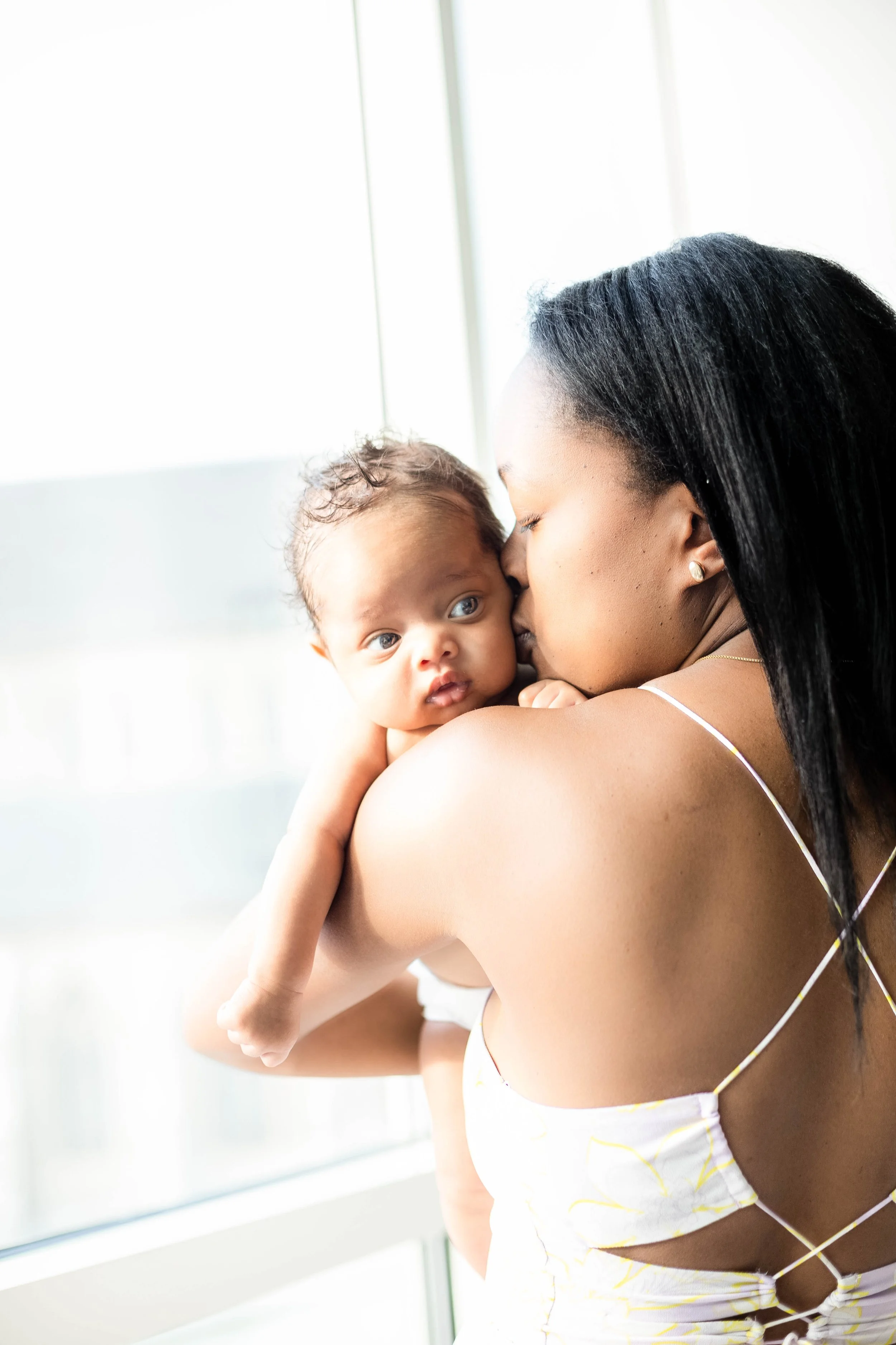 A woman with long black hair embraces and kisses a baby with curly hair and light brown skin, near a window with bright light in the background.