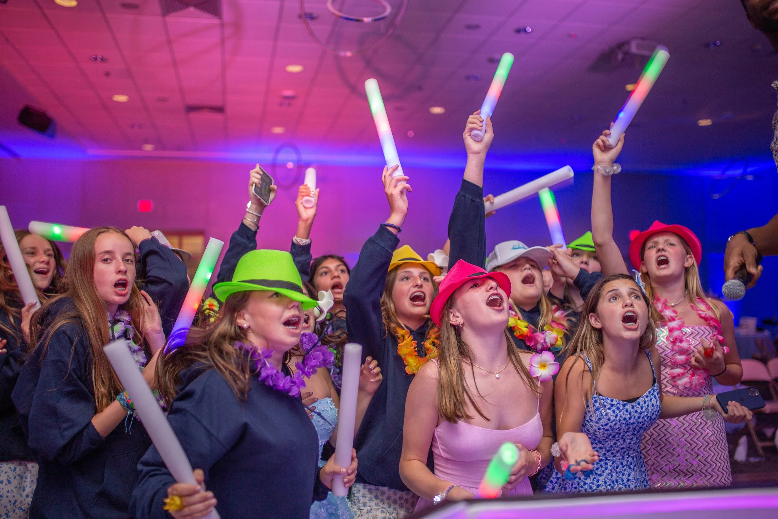 Group of young girls singing and dancing at a lively party or celebration, holding glow sticks and wearing colorful hats and accessories.