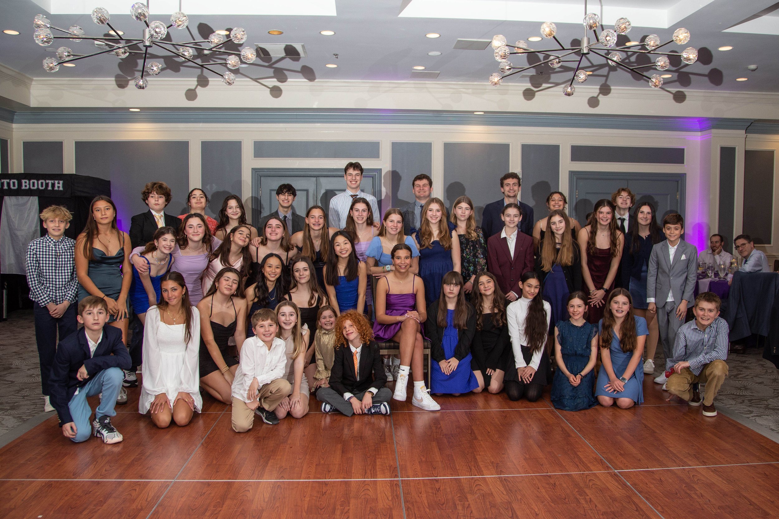 Group of children and teenagers dressed in formal attire posing for a group photo at an indoor event, with a ballroom or banquet hall setting.
