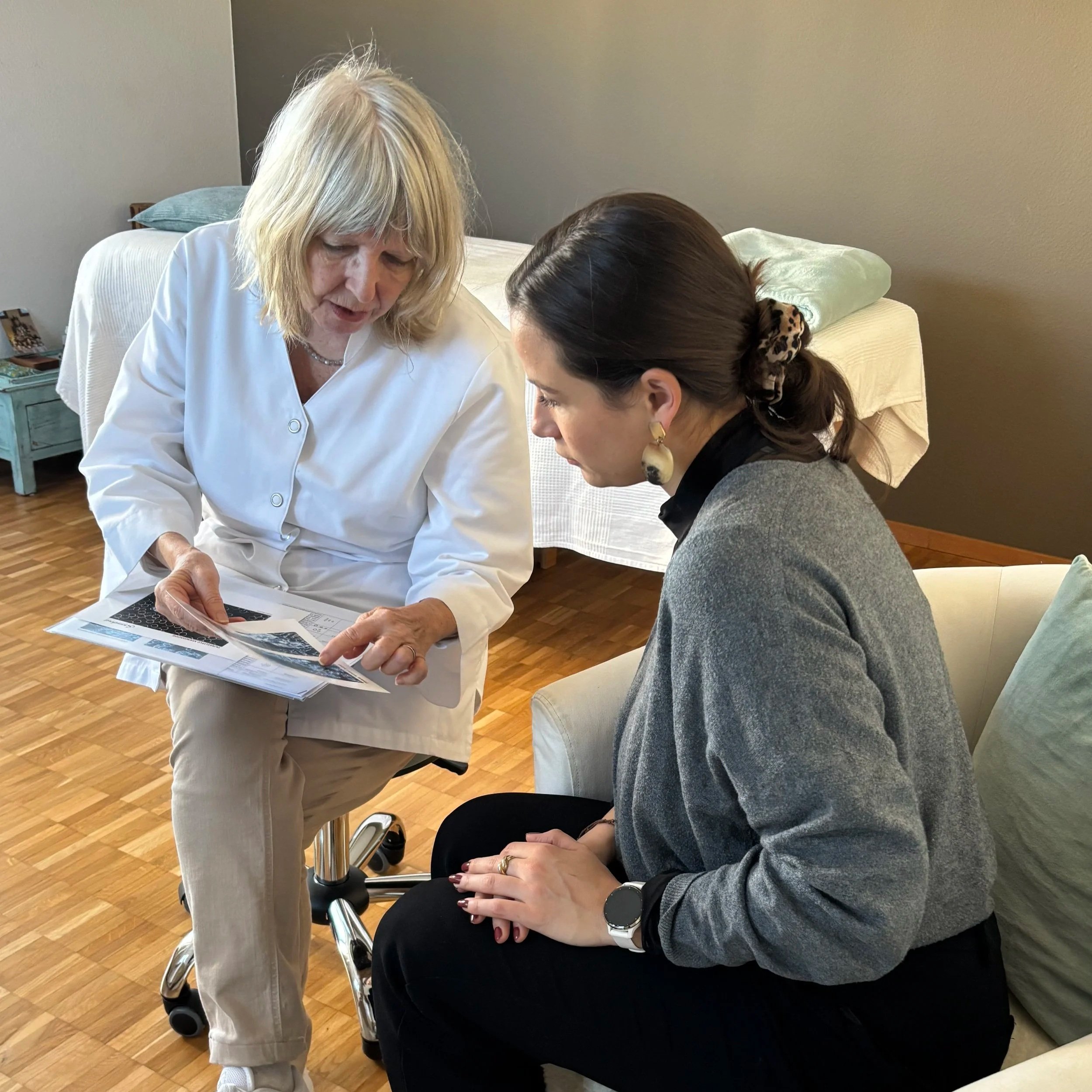 Zwei Frauen, eine ältere Ärztin und eine jüngere Patientin, unterhalten sich im Krankenzimmer, die Ärztin zeigt der Patientin einen medizinischen Bericht oder eine Bildgebung.