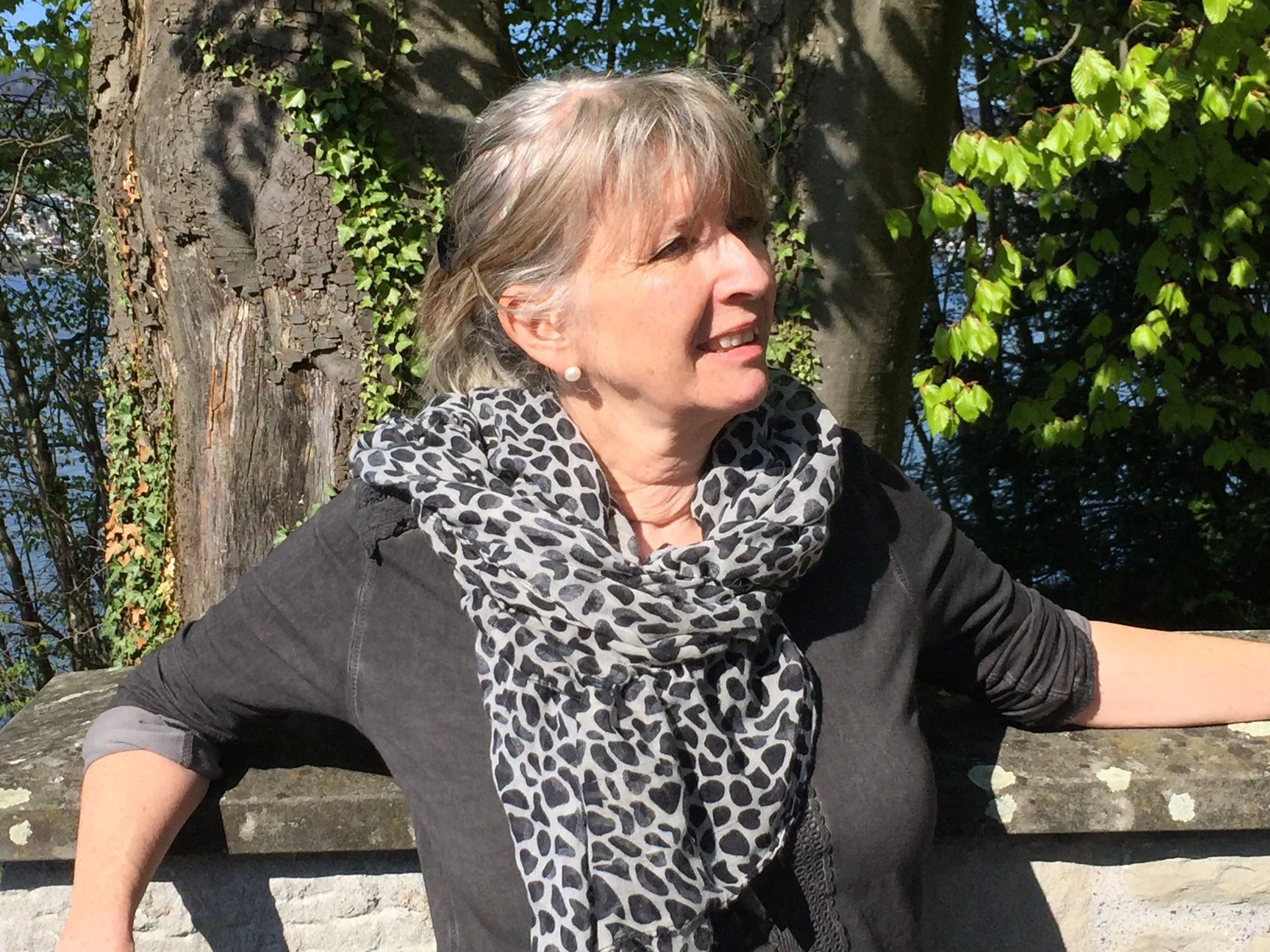 Eine älter Frau mit grauem Haar, trägt einen schwarzen Pullover, eine schwarz-weiß gepunktete Schal und Perlenohrringe, steht vor einem Baum und grünem Laub im Sonnenlicht.