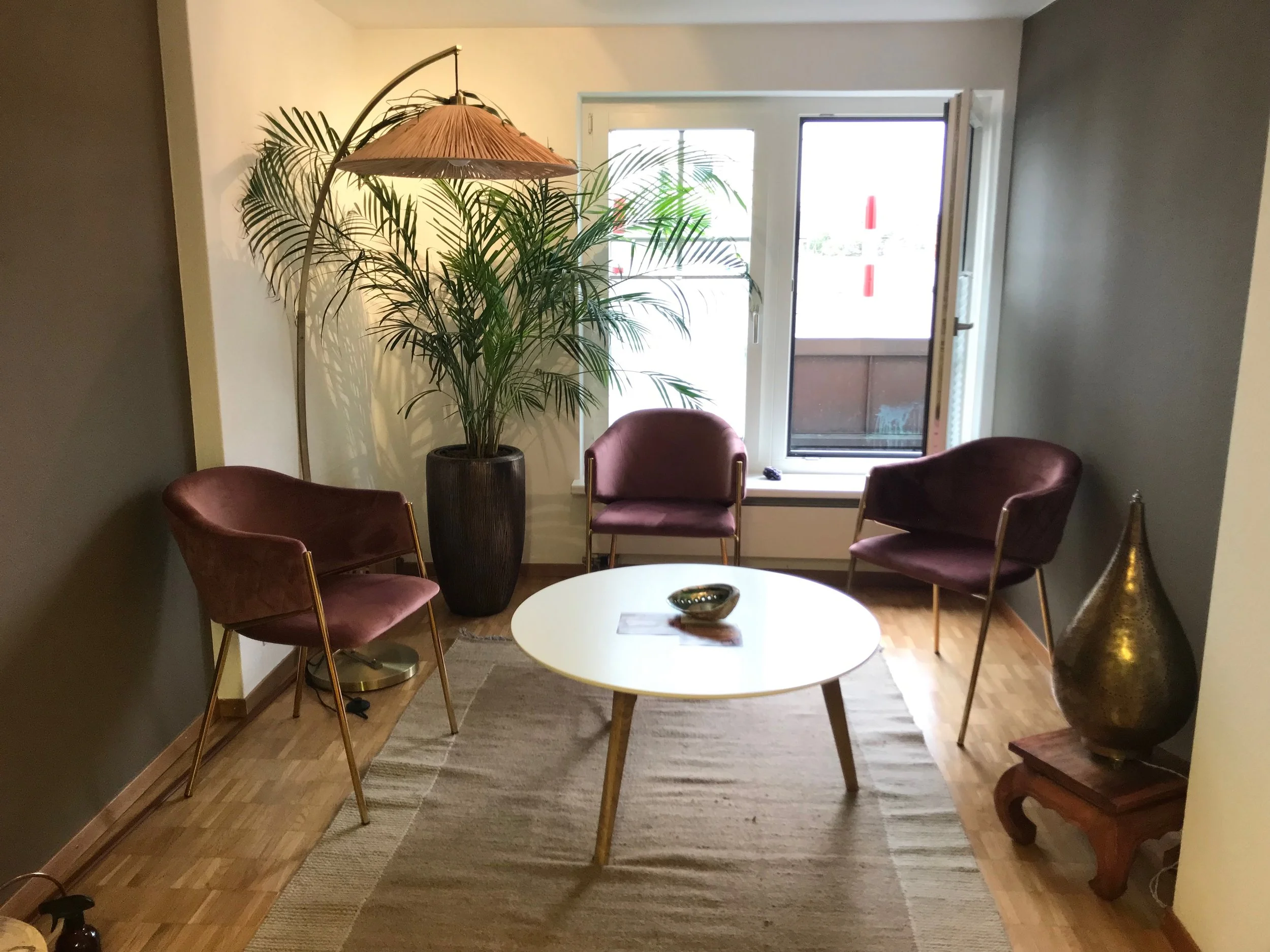 Gemütlicher Sitzbereich mit drei Stühlen um einen weißen runden Tisch, große grüne Pflanze in einer schwarzen Vase, ein Stehlampenschirm, ein großer goldener Dekorationsgegenstand auf einem kleinen Holzständer, eine offene Balkontür mit Blick nach draußen.