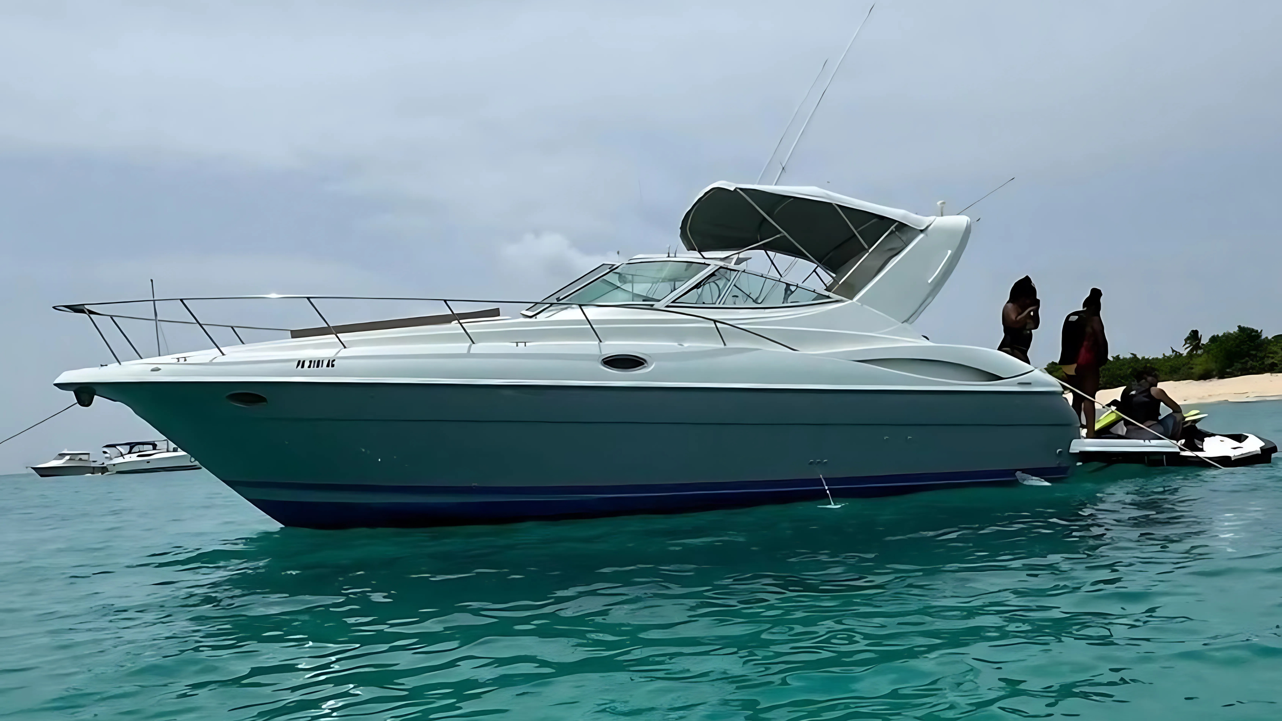 A white and teal yacht anchored near a beach with people standing and sitting on the boat, with another boat visible in the background.