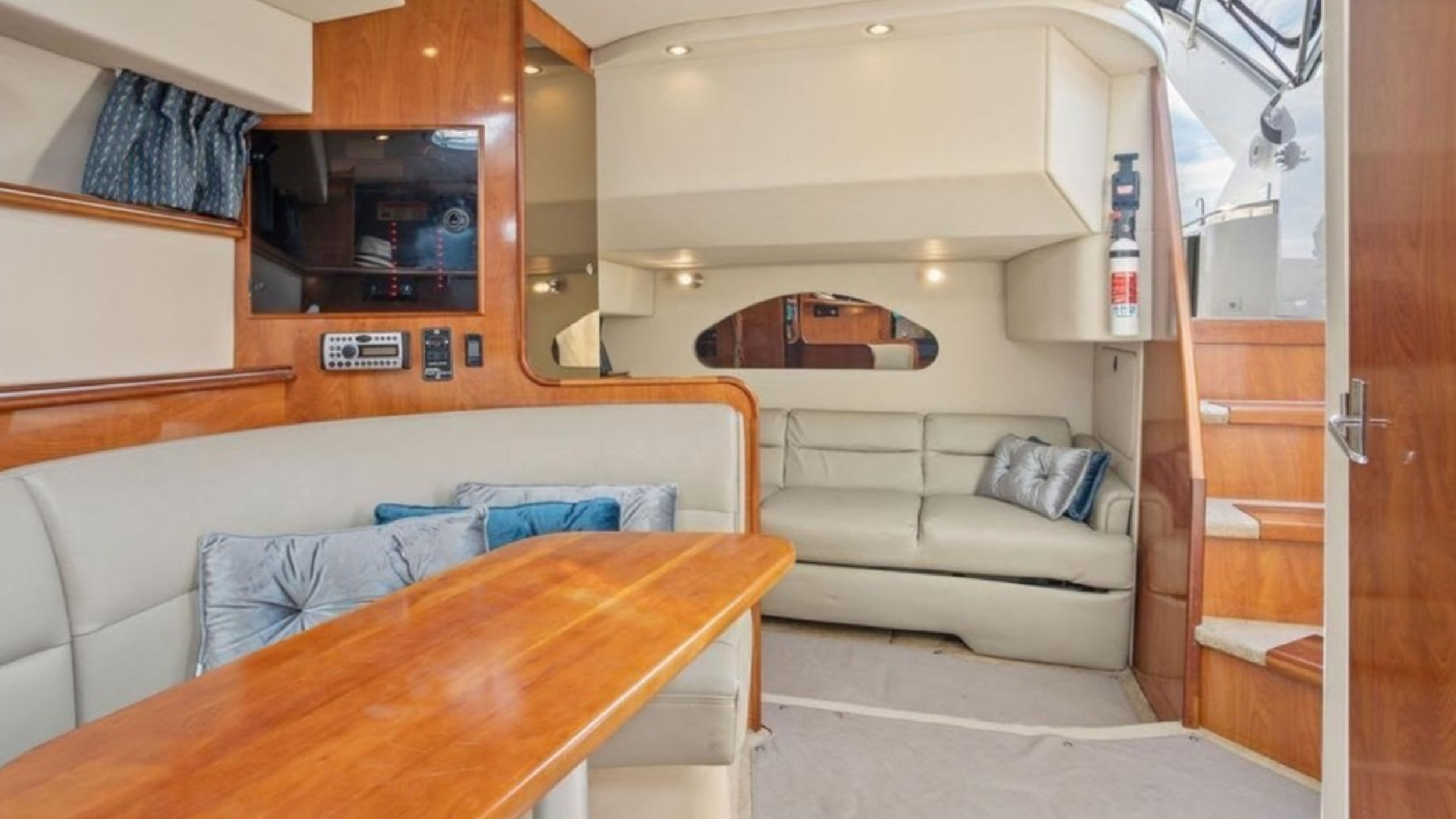 Interior of a boat or yacht with a dining area and sitting space, featuring beige seating, wooden paneling, and a staircase leading upstairs.