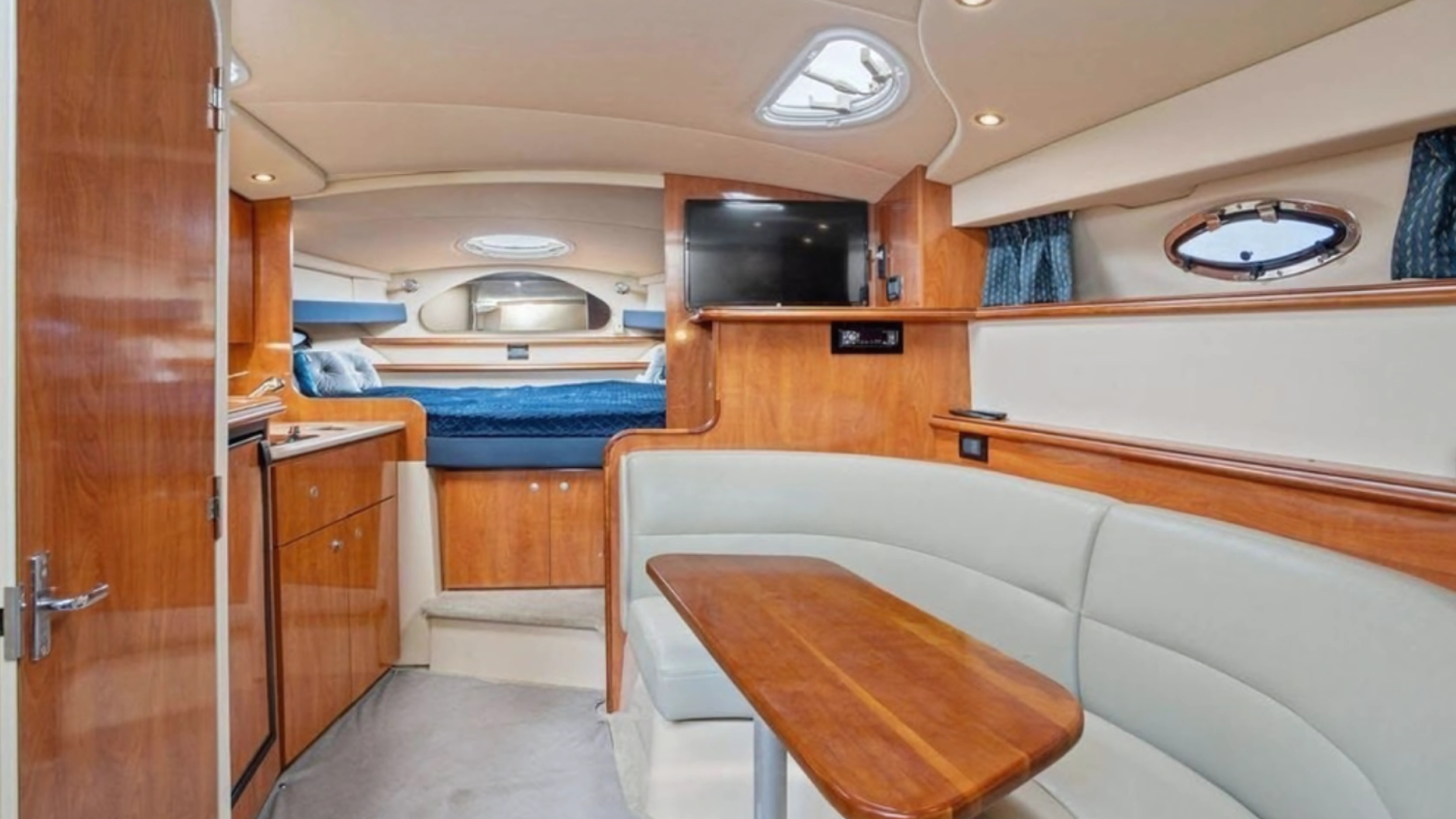 Interior of a boat or yacht cabin with a seating area, wooden cabinets, a bed in the background, and a small television.