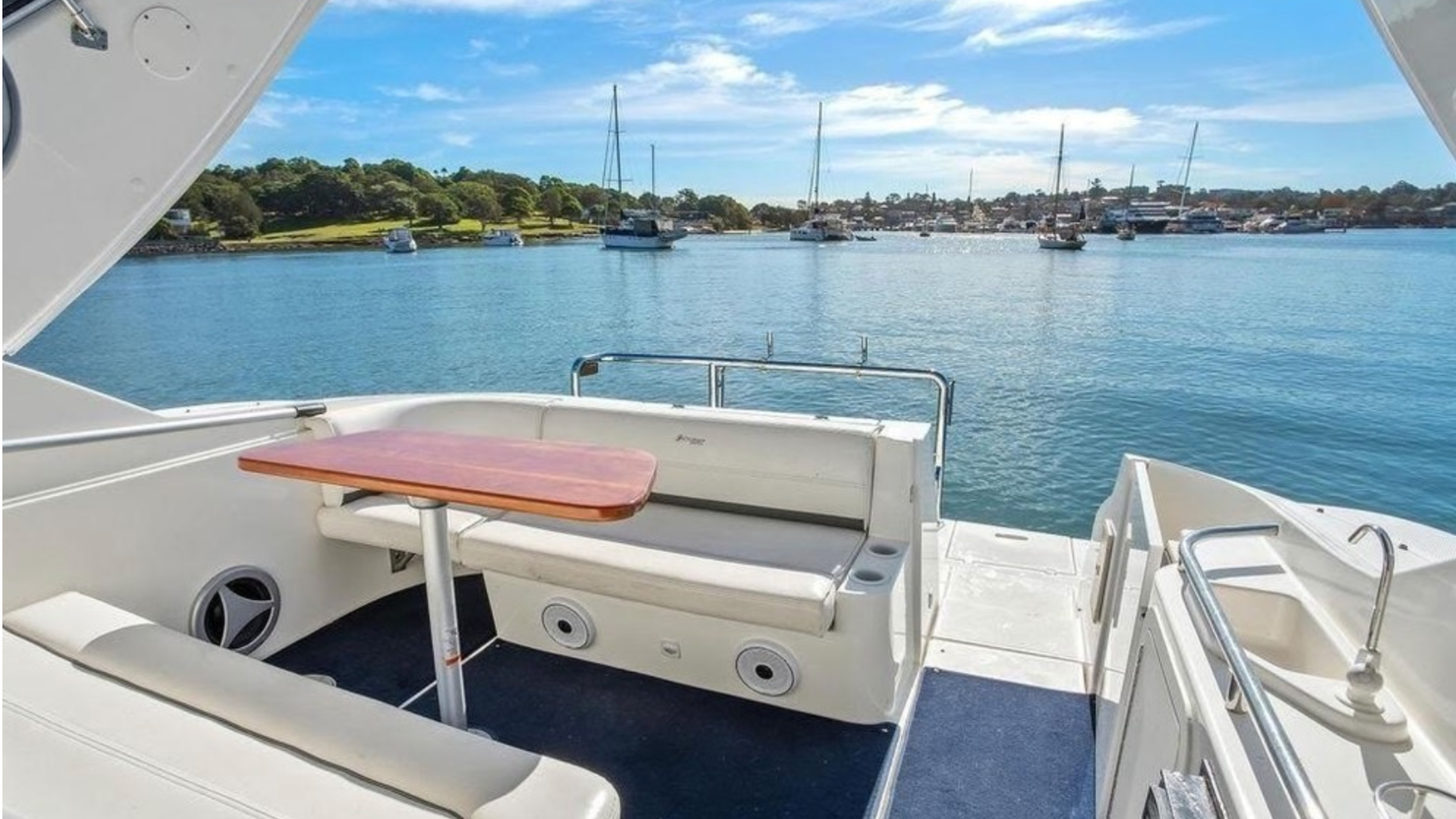 View from a boat showing a calm blue harbor with sailboats and yachts anchored, a green shoreline with trees, and a partly cloudy sky.