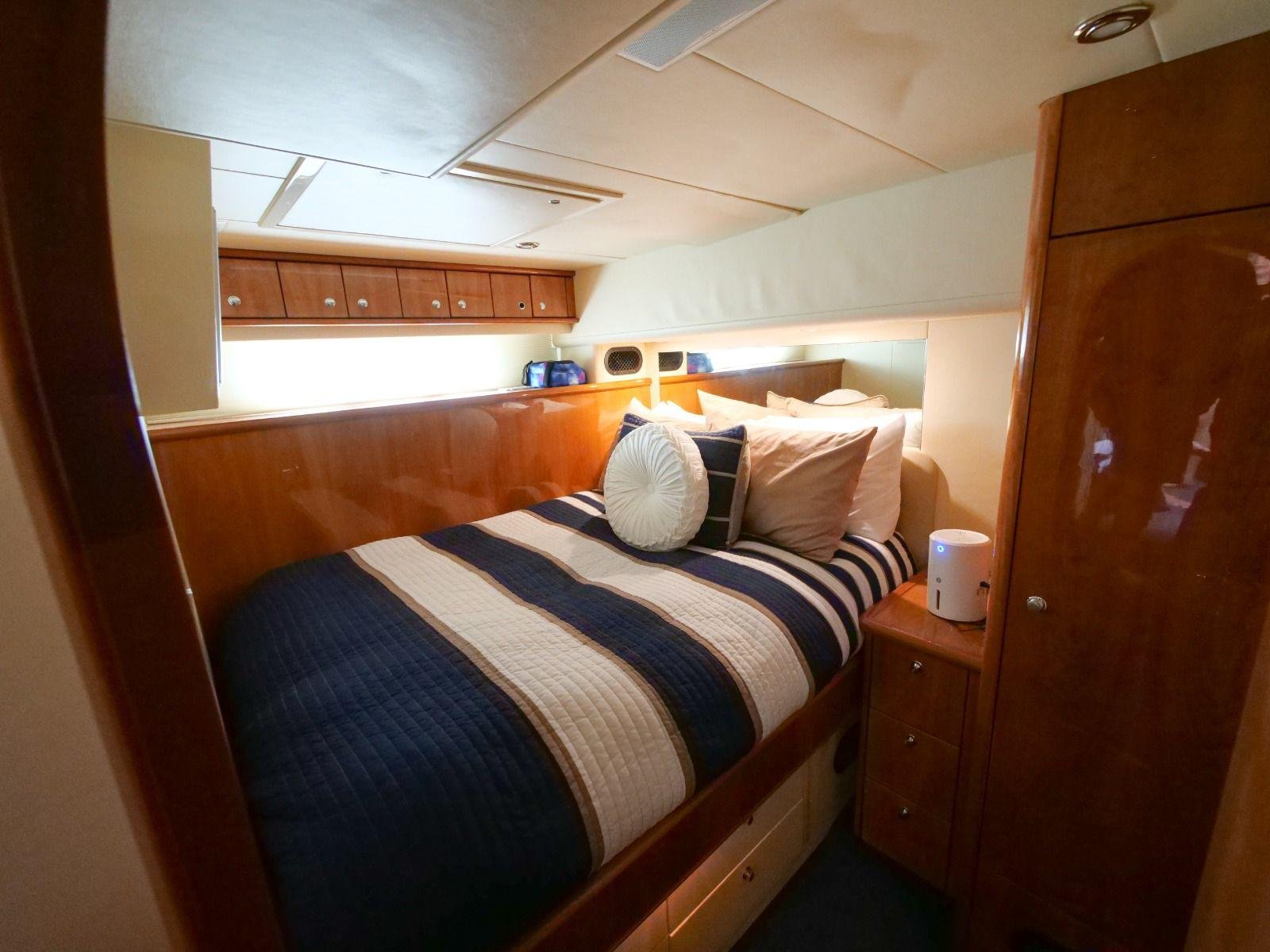 Small bedroom with a bed covered in a blue, white, and beige striped quilt, multiple pillows, and a wooden headboard. There is a wooden cabinet with drawers next to the bed, a small white device on top, and a narrow window with a limited view.
