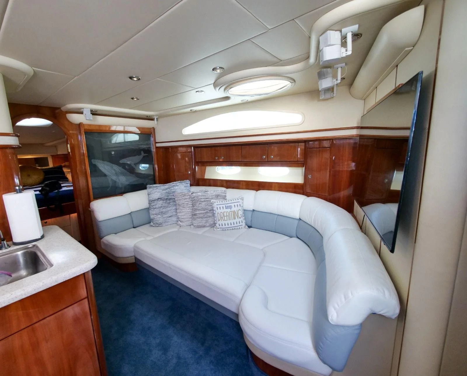 Living area inside a boat with a curved white leather sofa, throw pillows, a wooden wall with a window, and a wall-mounted flat-screen TV.