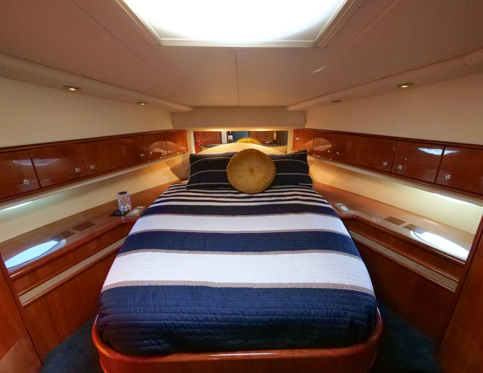Interior of a yacht bedroom with a neatly made bed, striped quilt, round pillow, built-in wooden cabinets along the sides, and a skylight overhead.
