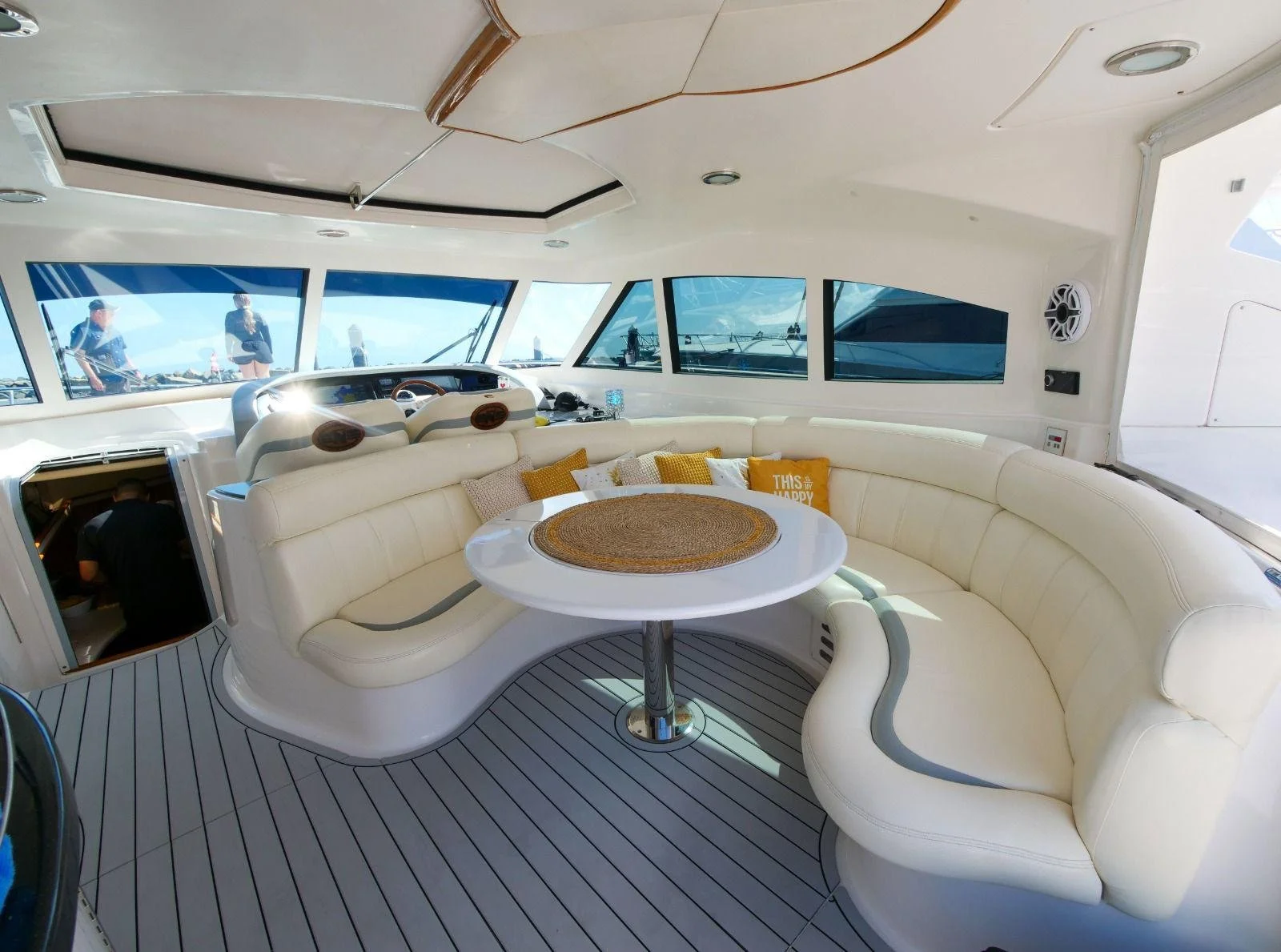 Interior of a yacht with a curved white leather seating area, a round table with a woven placemat, decorative pillows, large windows, and a view of the marina outside.