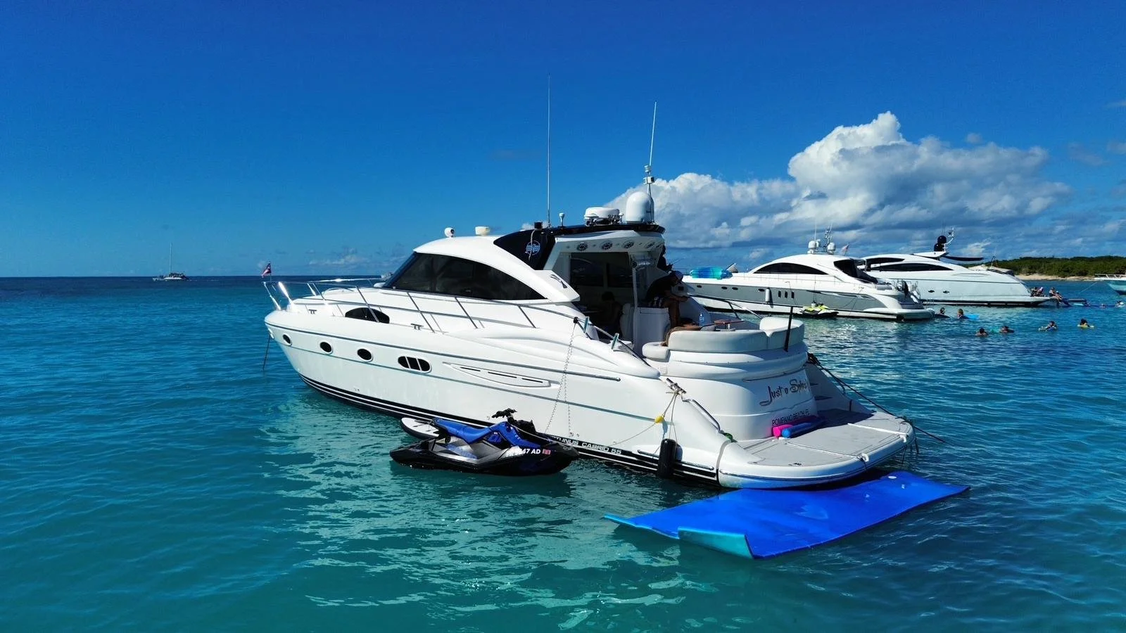 Several white yachts anchored on calm blue water under a partly cloudy sky, with people swimming nearby and a small blue floatation mat at the stern of the closest yacht.