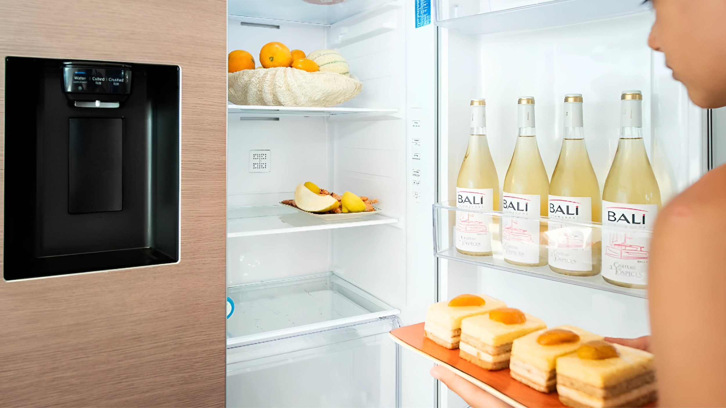 Open refrigerator with fruit, wine bottles, and a person placing dessert inside.