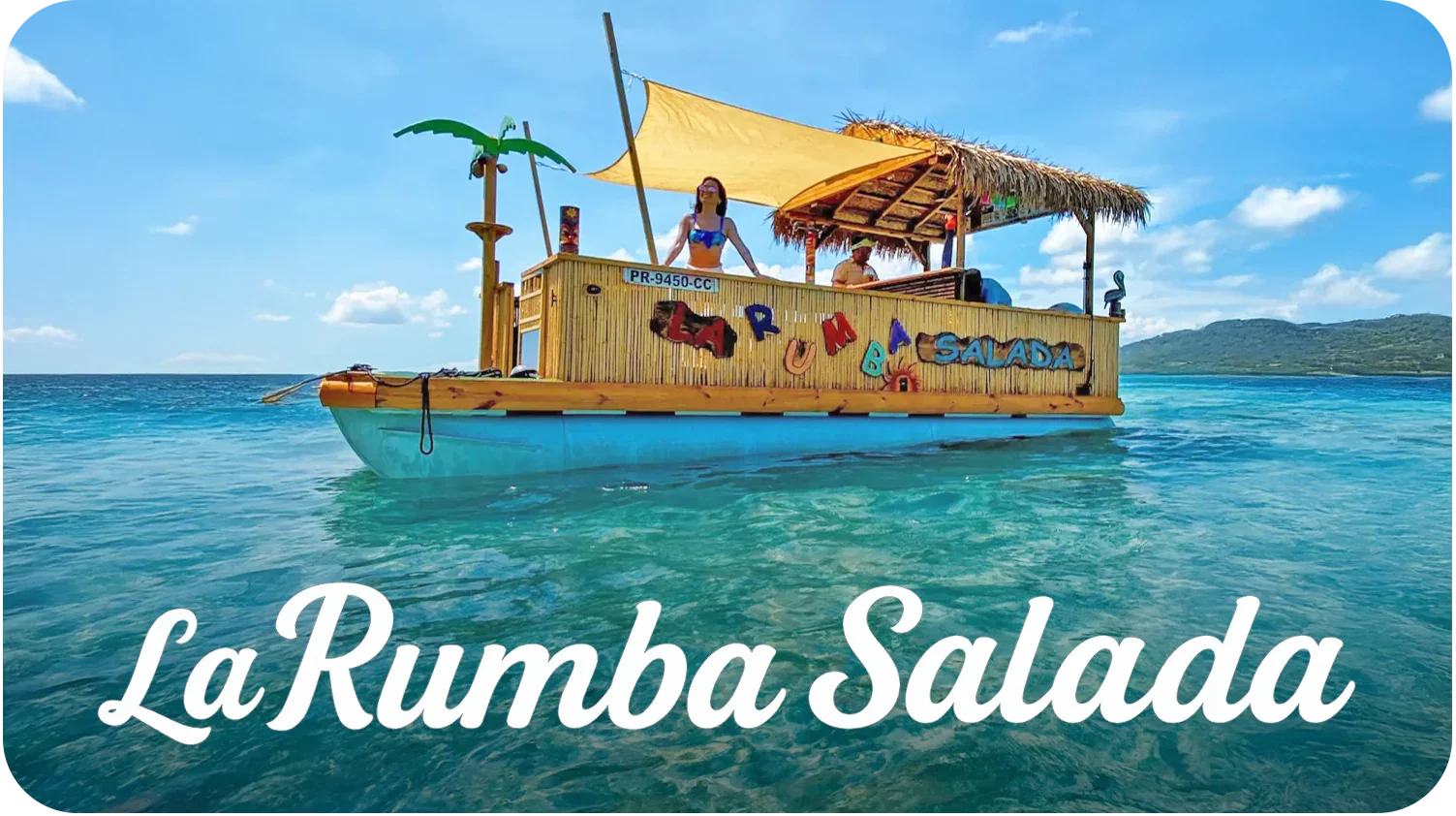 A floating bar named La Rumba Salada decorated with colorful letters, with a woman and man on board, on clear blue water with hills in the background, under a partly cloudy sky.