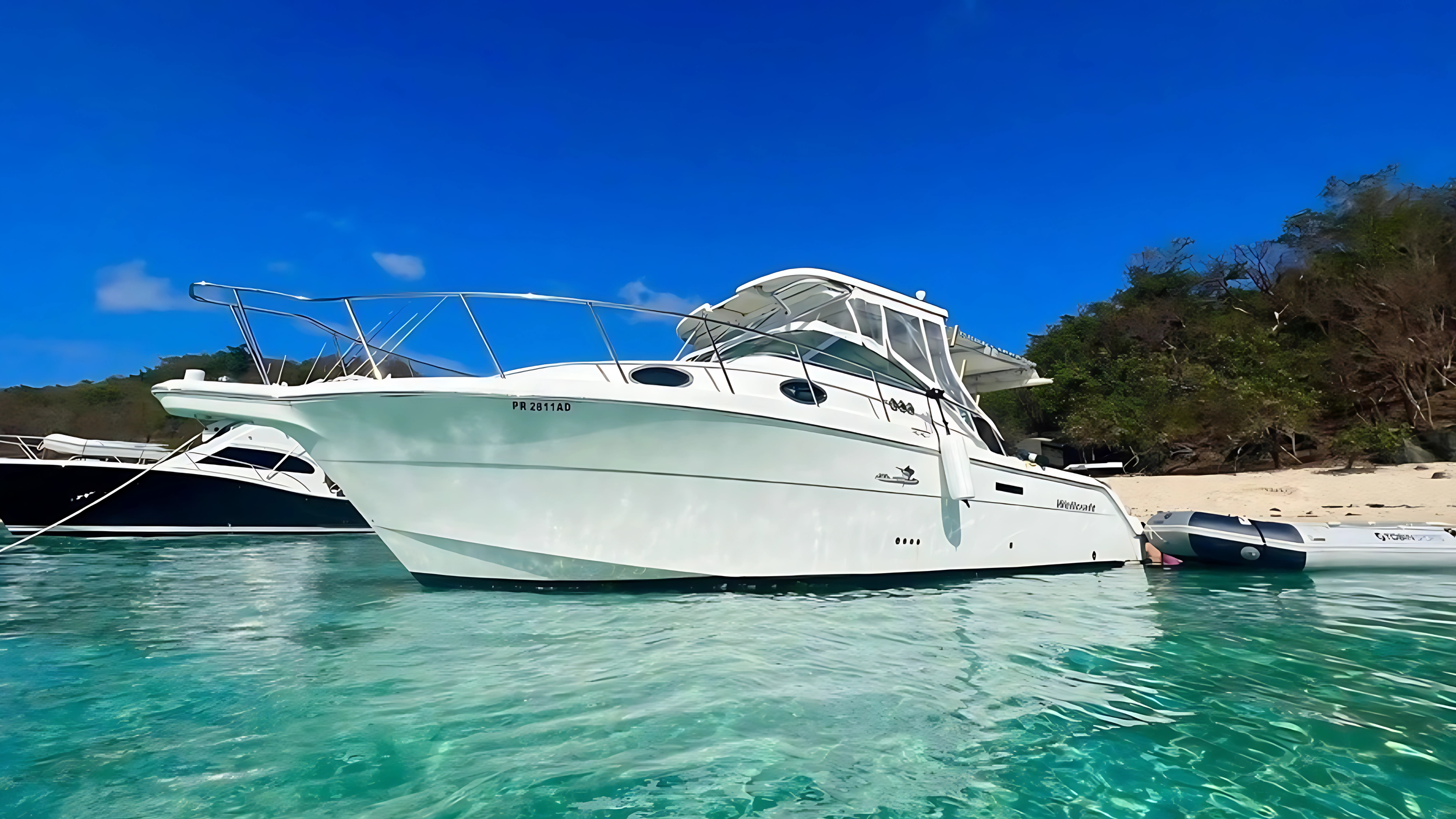 A white yacht anchored near a sandy beach with trees on a bright sunny day.