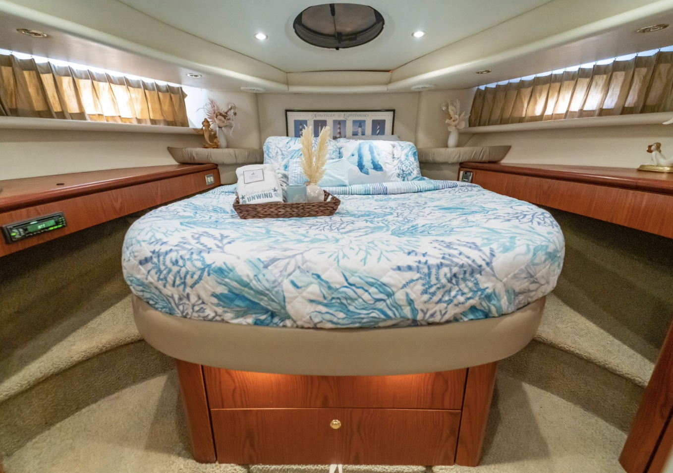 Luxury yacht bedroom interior with a round bed, nautical-themed bedding, and decorative pillows. Wooden cabinetry lines the room, and soft lighting emanates from above. Large porthole and curtains add a nautical touch.
