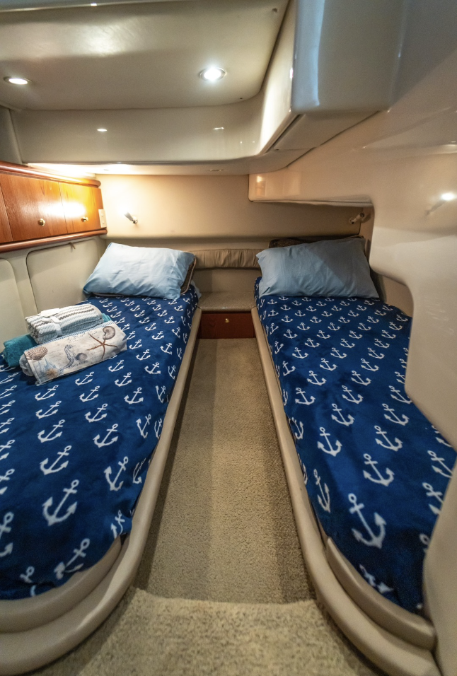 Boat cabin with twin beds, navy anchor-patterned bedding, blue pillows, towels on left bed, wooden cabinets, beige carpet, and overhead lighting.