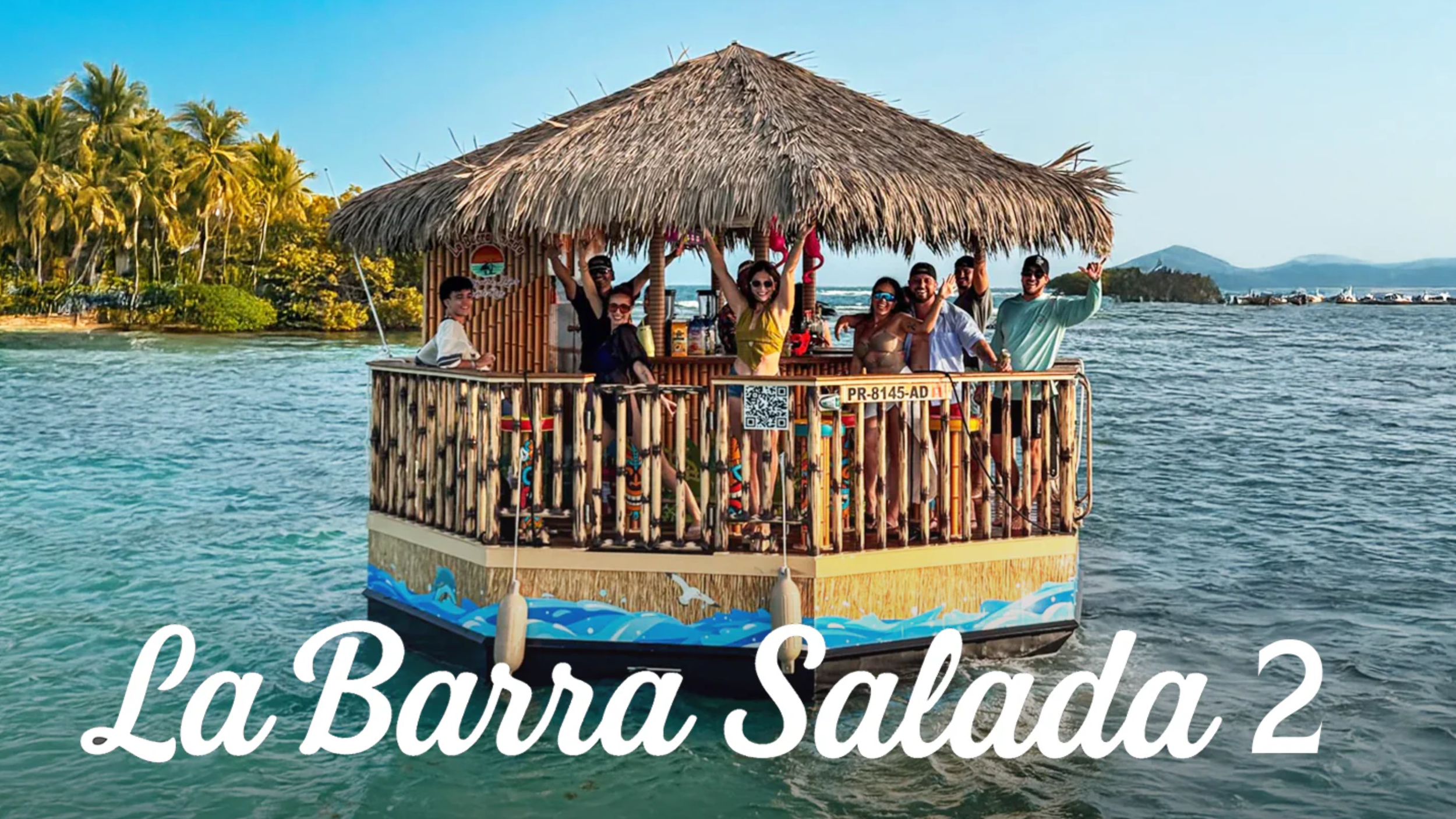 A group of people enjoying a boat ride on a floating bar with a thatched roof, on clear water near a tropical island with palm trees and a distant mountainous landscape.