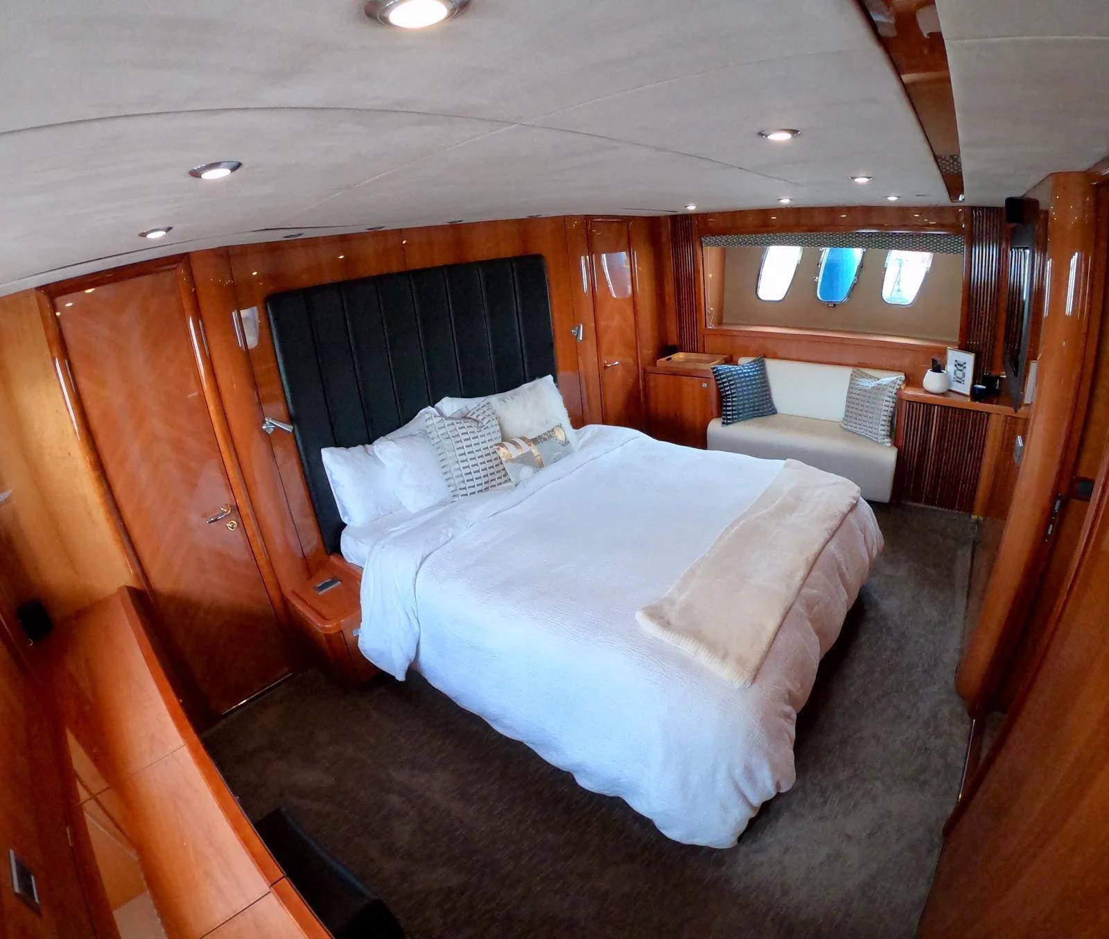 Luxurious yacht bedroom interior with wooden paneling, a large bed, plush pillows, compact sofa, and small table near window portholes.