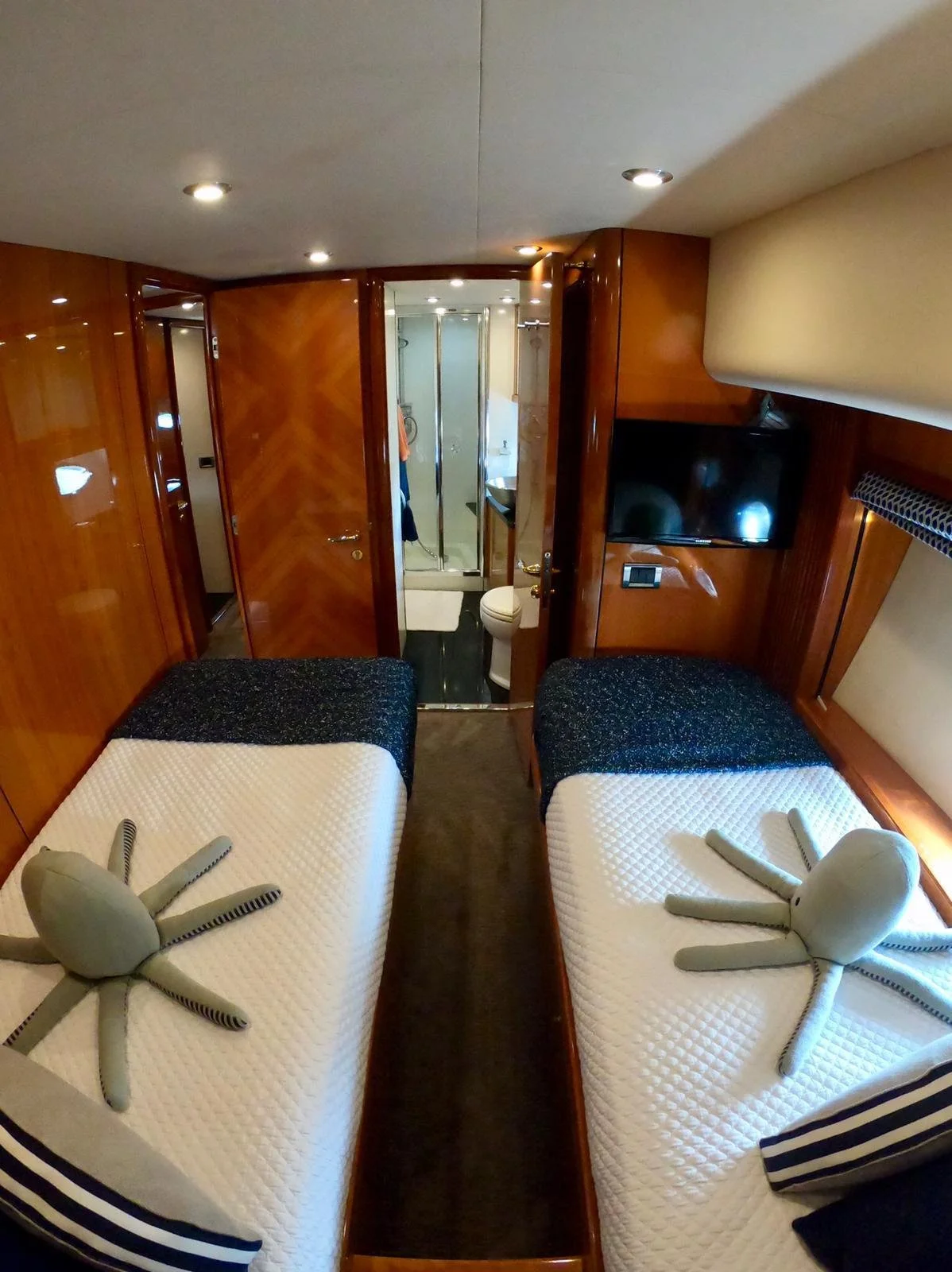 Luxury yacht cabin interior with two twin beds, plush octopus toys, striped pillows, wood paneling, mounted TV, and ensuite bathroom with shower.