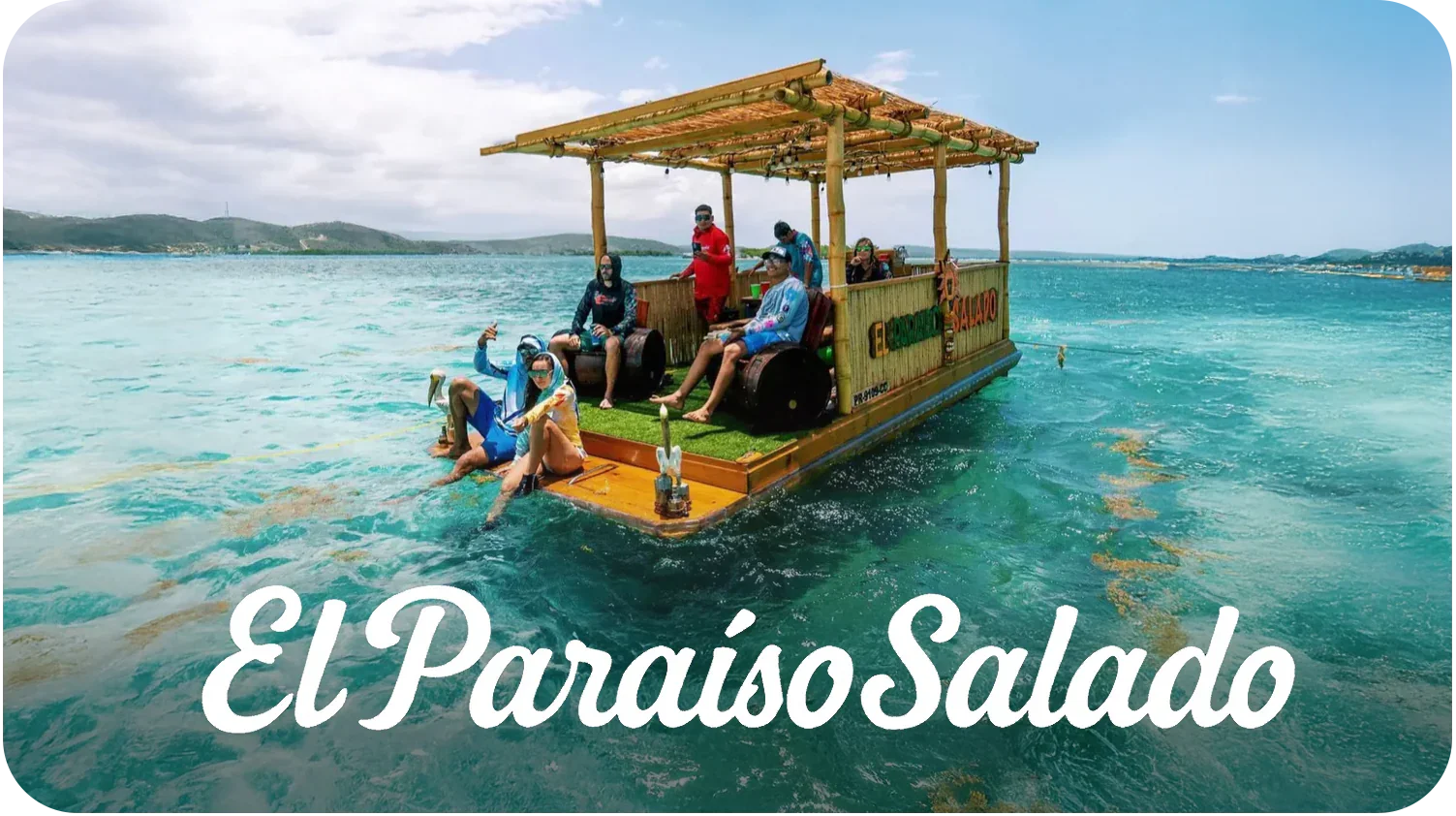 Group of people on a floating wooden raft with a thatched roof, decorated with signs, on turquoise water with a distant shoreline and mountains in the background.