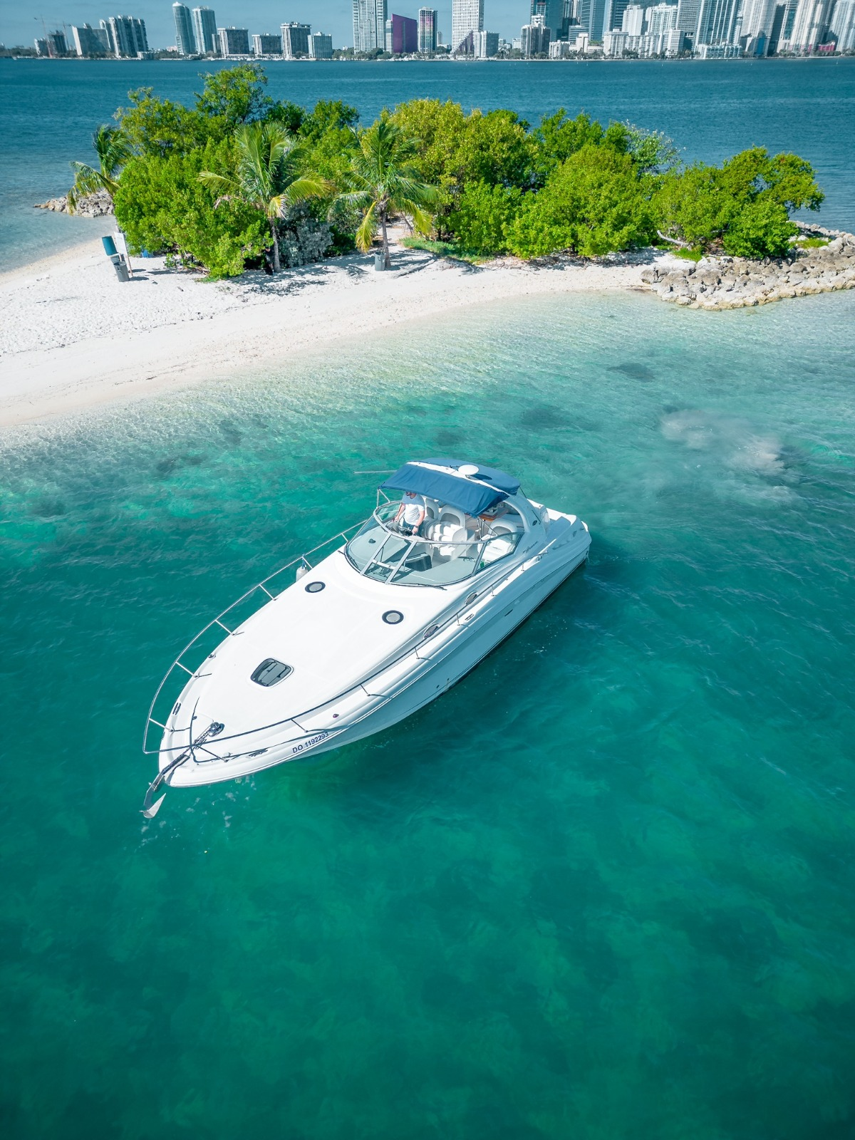 A white motorboat cruising near a sandy island with green trees and palms, with a city skyline in the background.