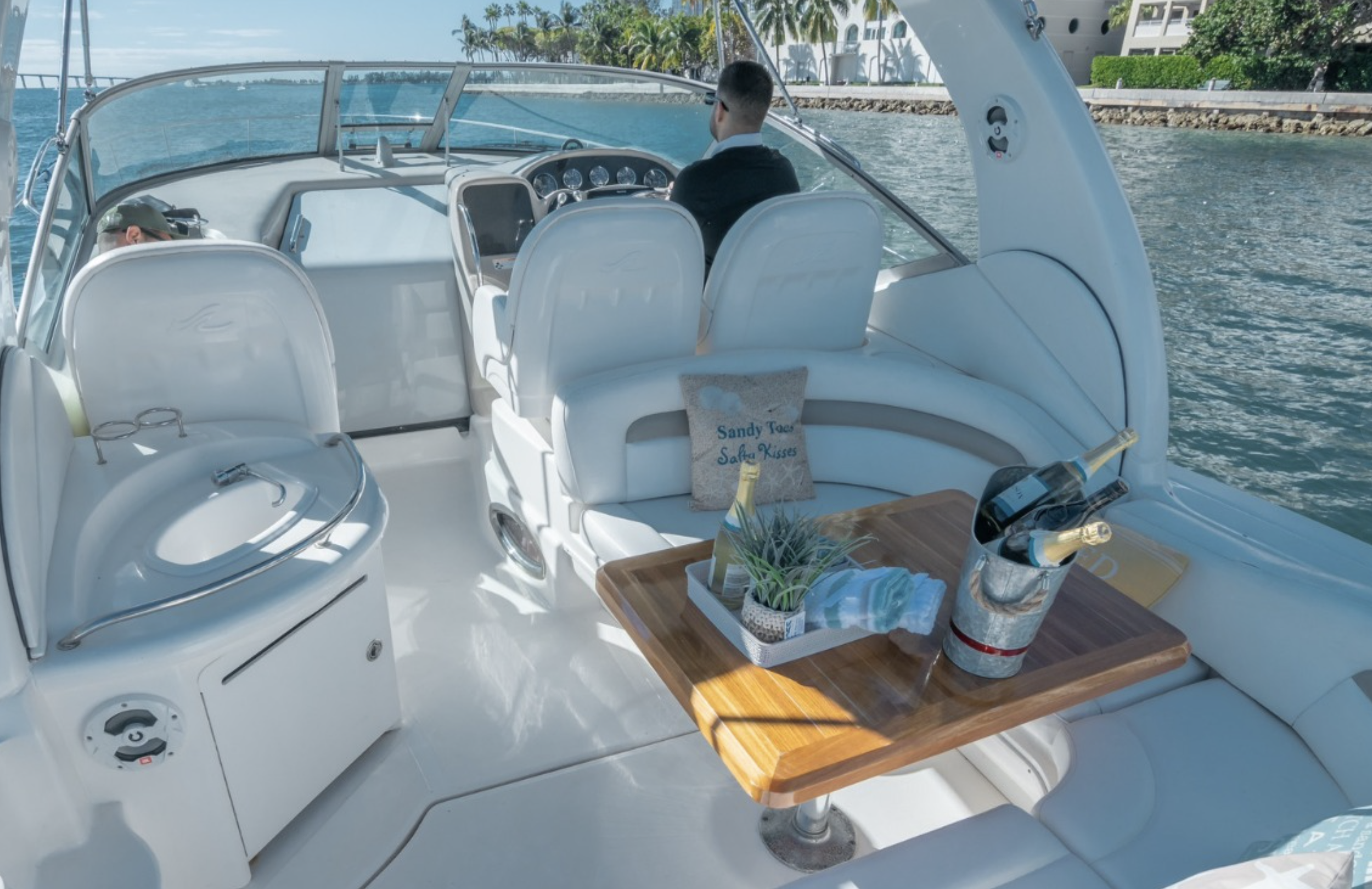 Interior of a luxury boat with white seating, a wooden table with wine bottles, a plant, and a decorative pillow that reads 'Sandy Toes, Salty Kisses.' Two people are seated at the front, one in sunglasses and the other facing away. The boat is on the water near a shoreline with palm trees and buildings.