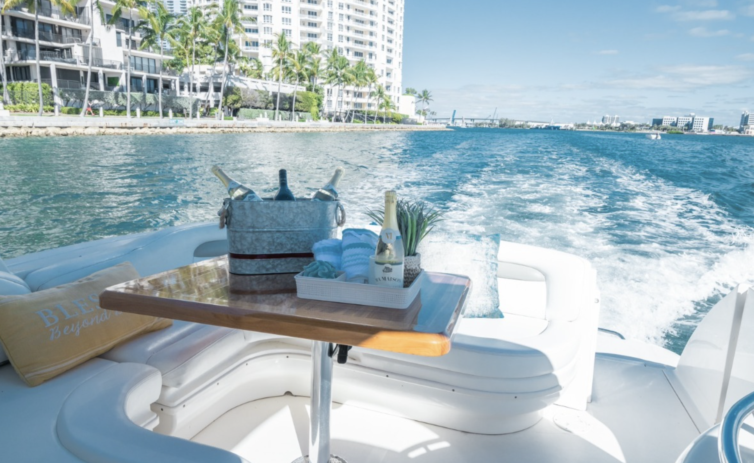 On a boat, a wooden table with beverage bottles in a cooler, a tray with a bottle of champagne, a small plant, and towels, with the water and buildings in the background.