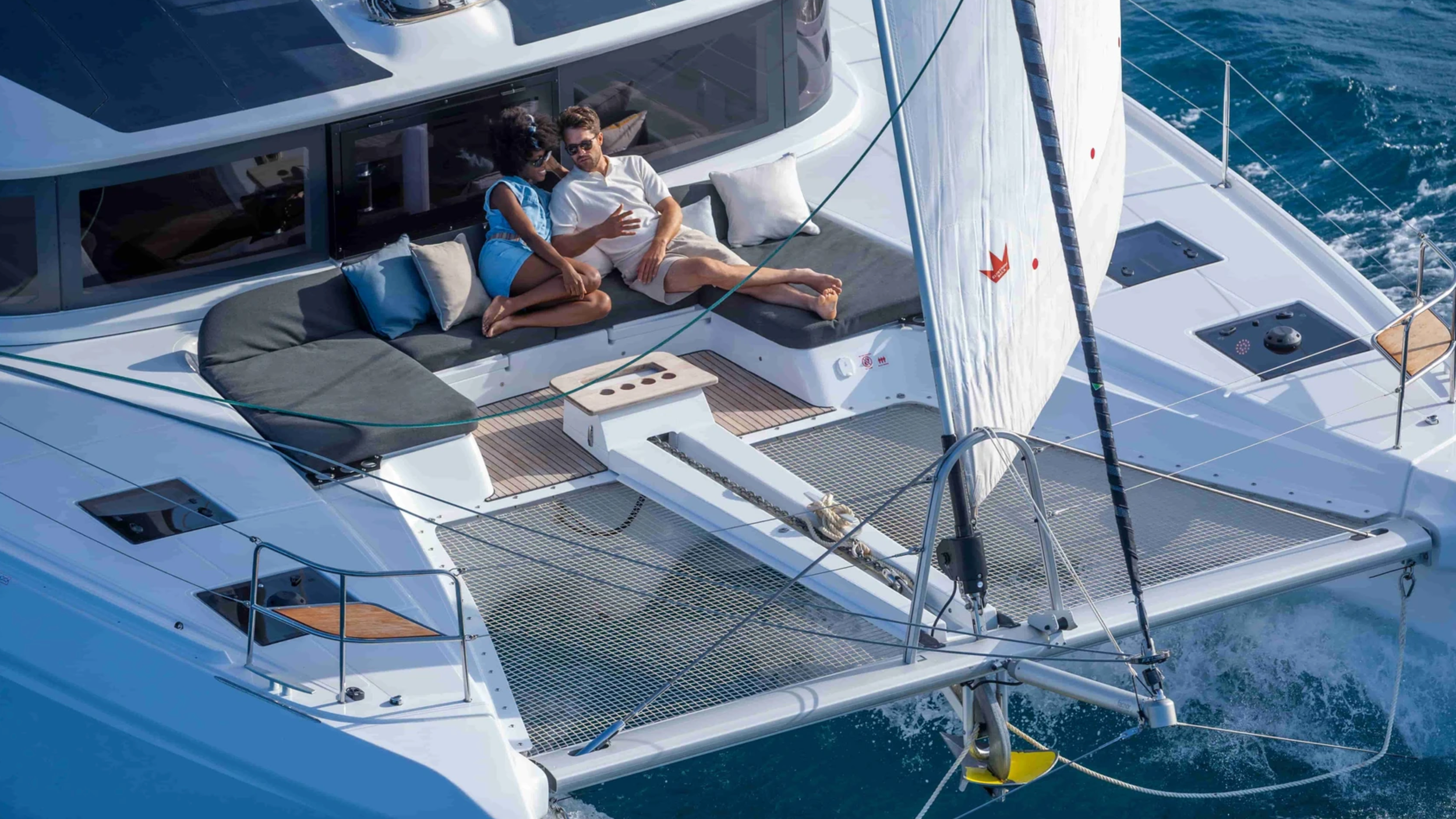 Couple relaxing on a catamaran, lounging on deck, open sea background.