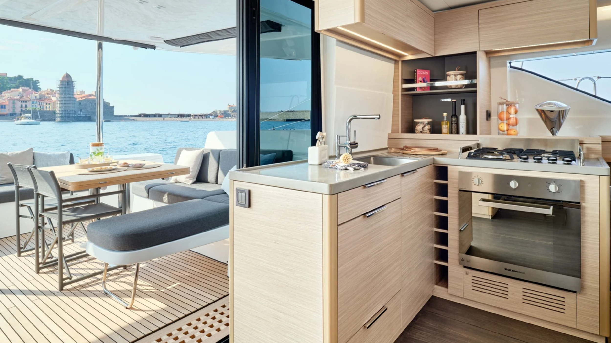 Modern yacht kitchen with light wood cabinets, stainless steel sink, and stove; open view to outdoor seating and ocean.