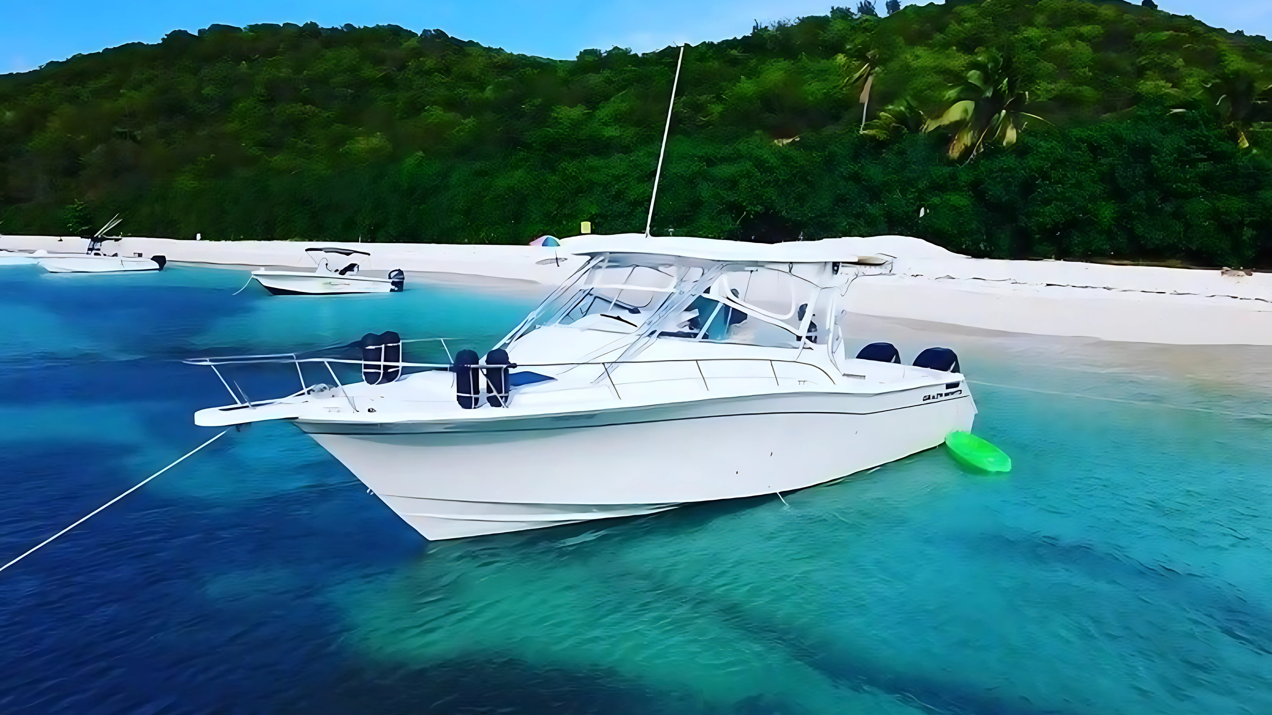 A white motor yacht docked in clear turquoise water near a white sandy beach with green trees and hills in the background.
