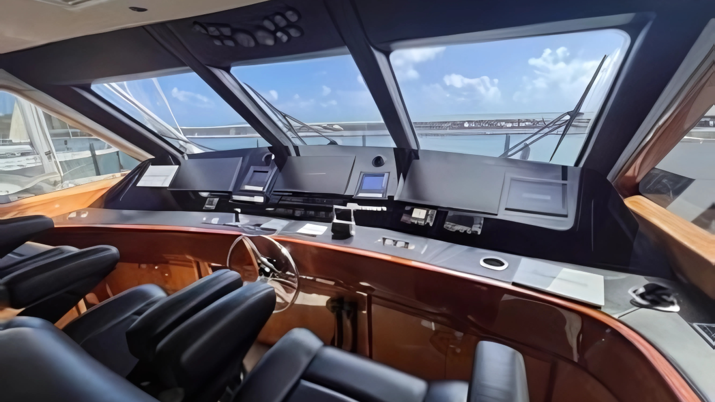 View of a boat cockpit with black seats, autopilot controls, and large windows showing the marina and blue sky outside.