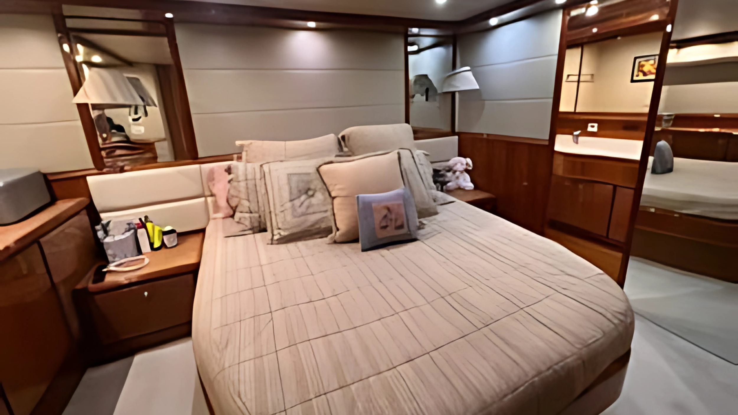 A cozy hotel room with a bed, multiple pillows, wall-mounted lamps, a small side table with a towel, and wooden decor. In the background, a partial view of another room or area with a bed.