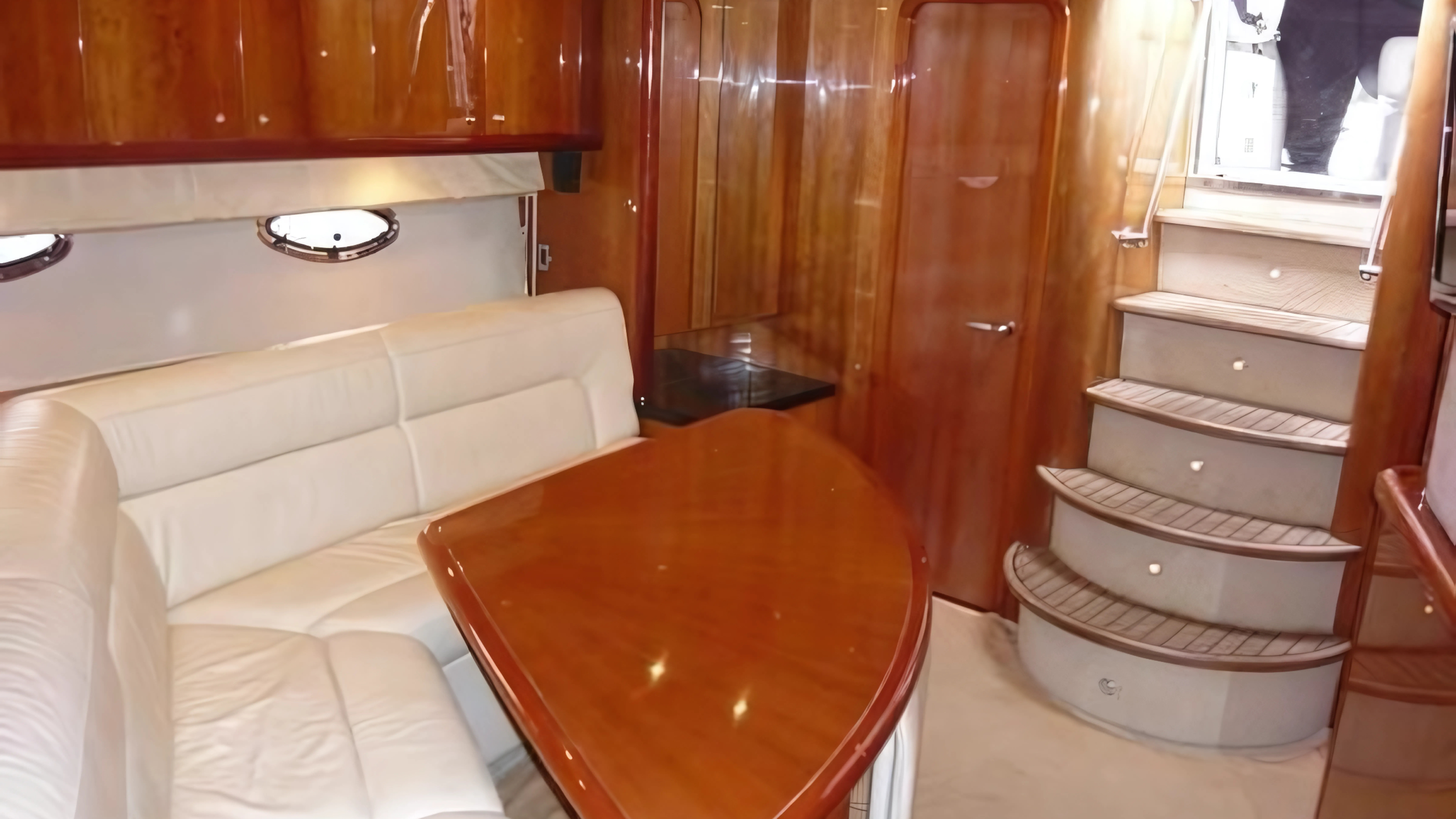 Luxury yacht interior with white leather seating, polished wooden table, and stairs leading to an upper deck.