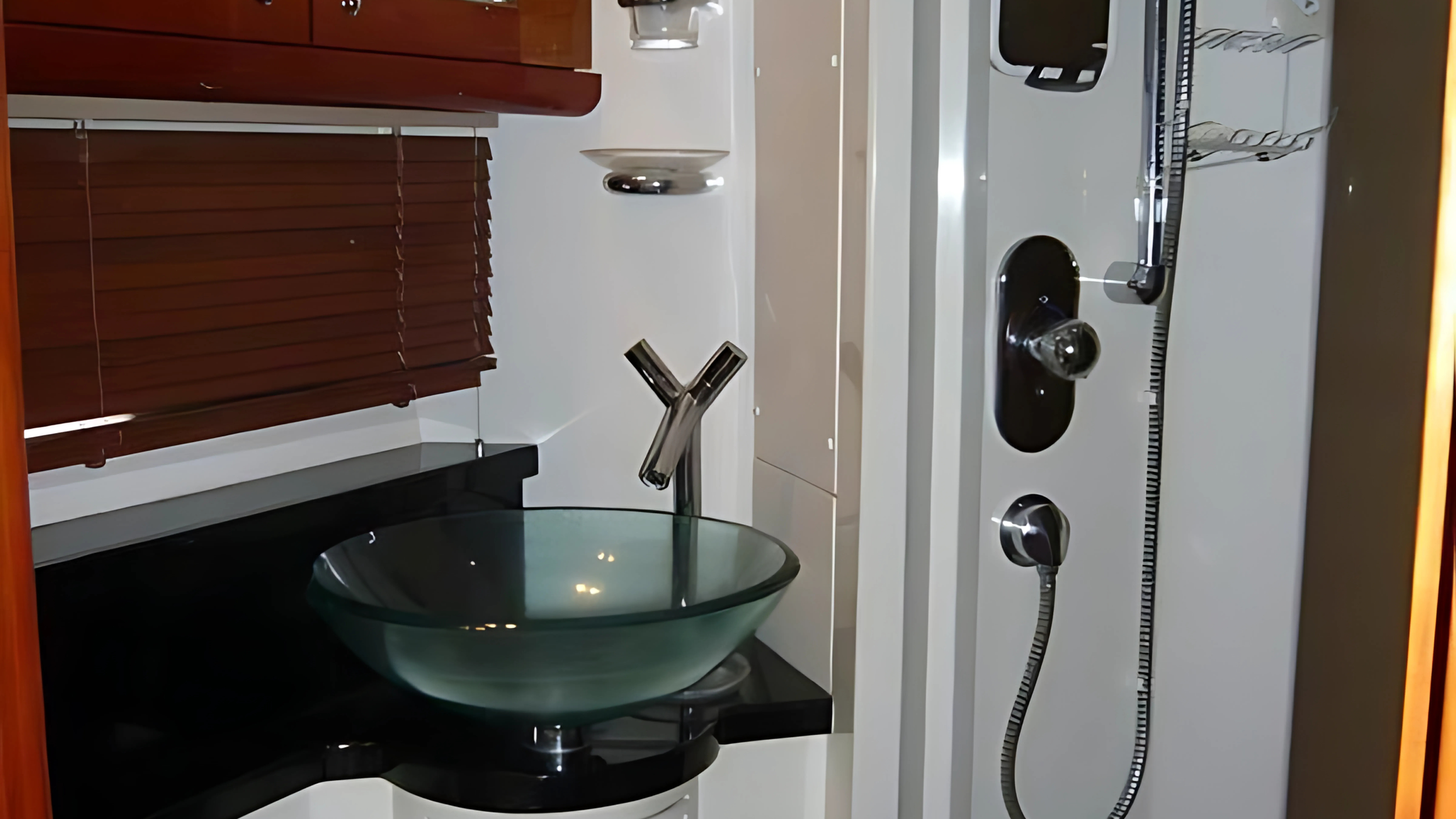 Modern bathroom with glass sink, chrome faucet, shower panel, wooden blinds, and black countertop.
