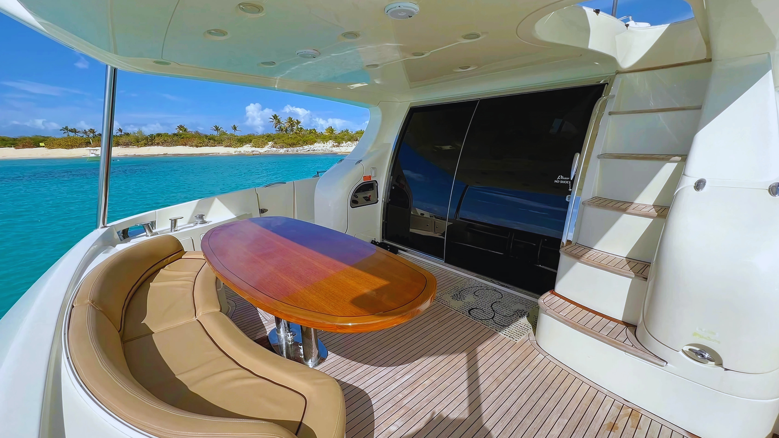 Luxury yacht deck with table and seating, view of turquoise ocean and sandy beach with palm trees.