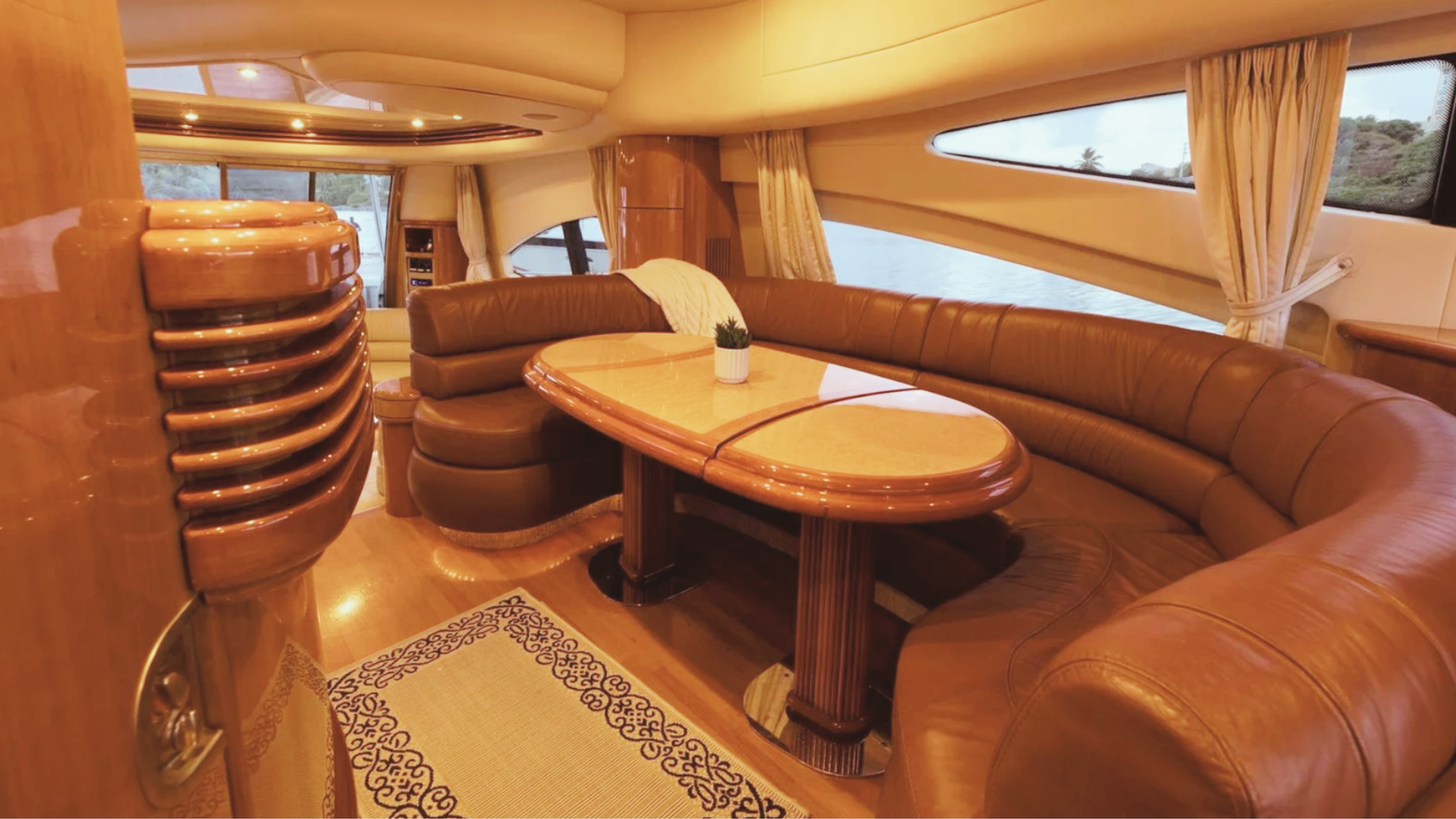 Luxurious yacht interior with leather seating, wooden table, decorative plant, and panoramic windows.