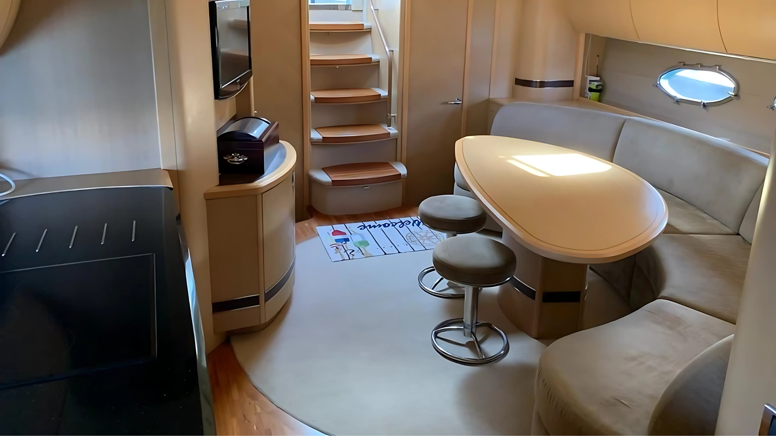 Interior of a yacht with a modern dining area, beige curved seating, an oval table, bar stools, wooden steps, and a small window.