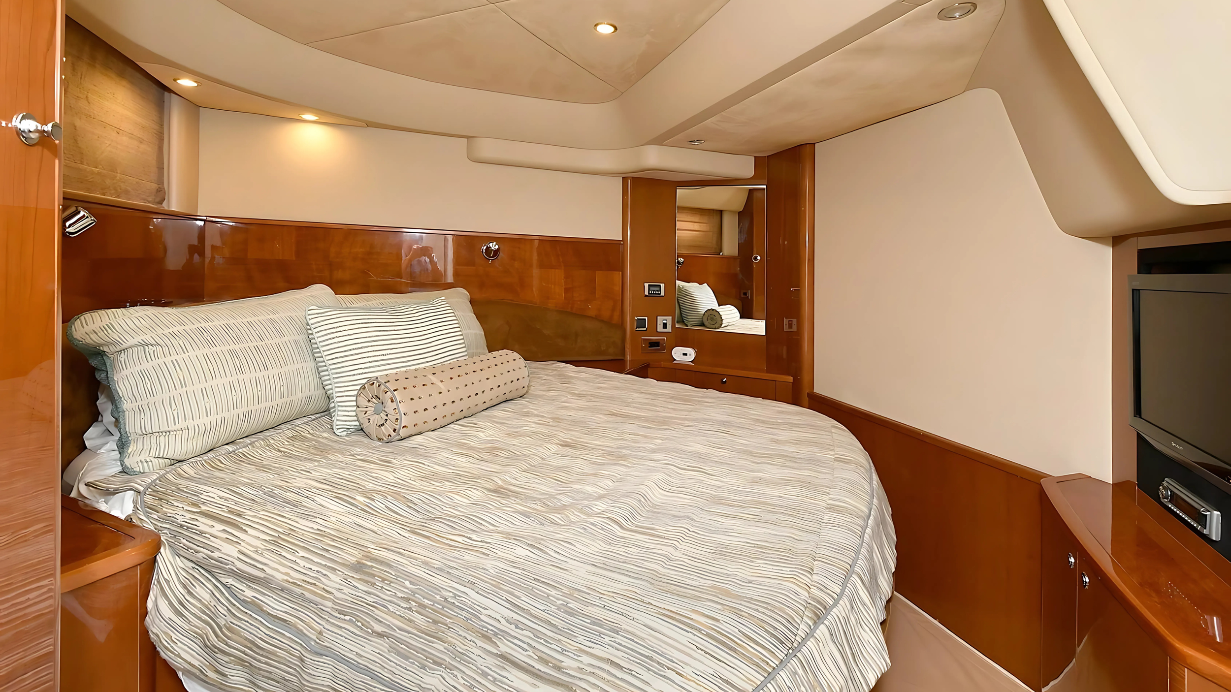 Luxury yacht cabin interior with a bed, wood paneling, built-in TV, bedding, mirrors, and ambient lighting.