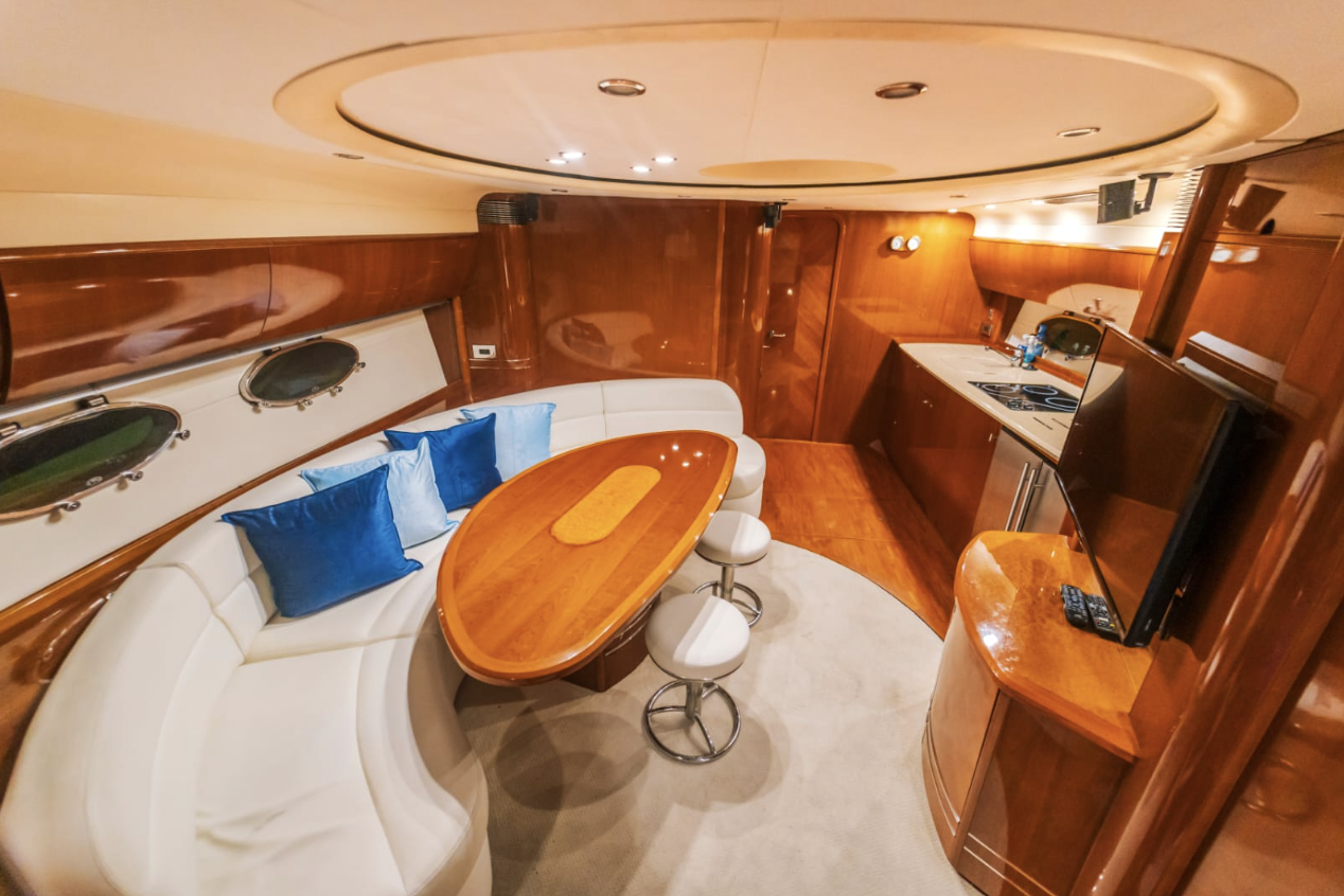 Interior of a yacht with a curved white sofa with blue pillows, a wooden dining table with two white stools, a small kitchen area with a stovetop, and a TV on a wooden stand.