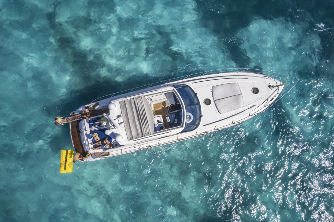 From a bird's eye view, a white yacht is floating on clear blue water with several people on board, some sitting and some standing near the edge.