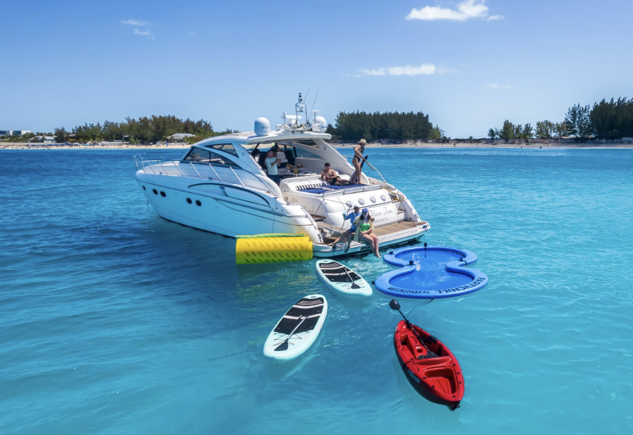 A group of people on a white yacht anchored in turquoise water near a shoreline with trees, with paddleboards, a kayak, and an inflatable float nearby.